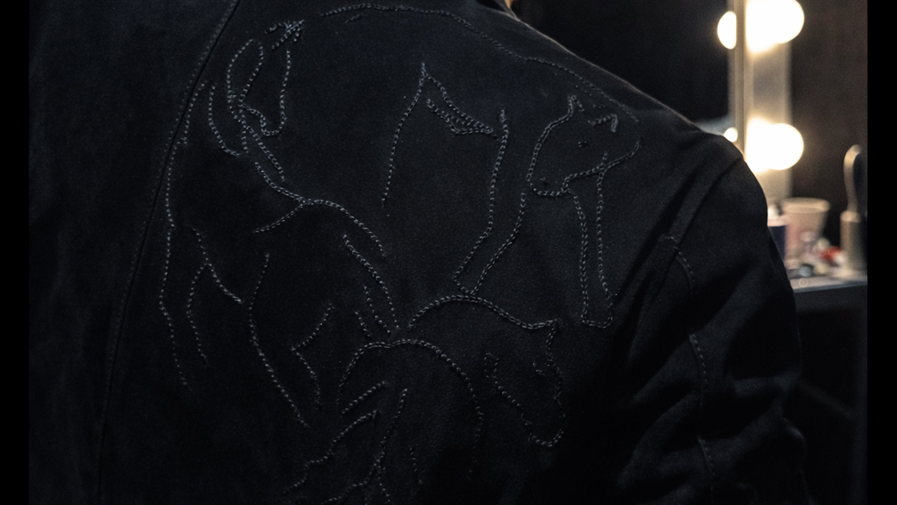 Person wearing a dark jacket with embroidered designs in a dimly lit room.