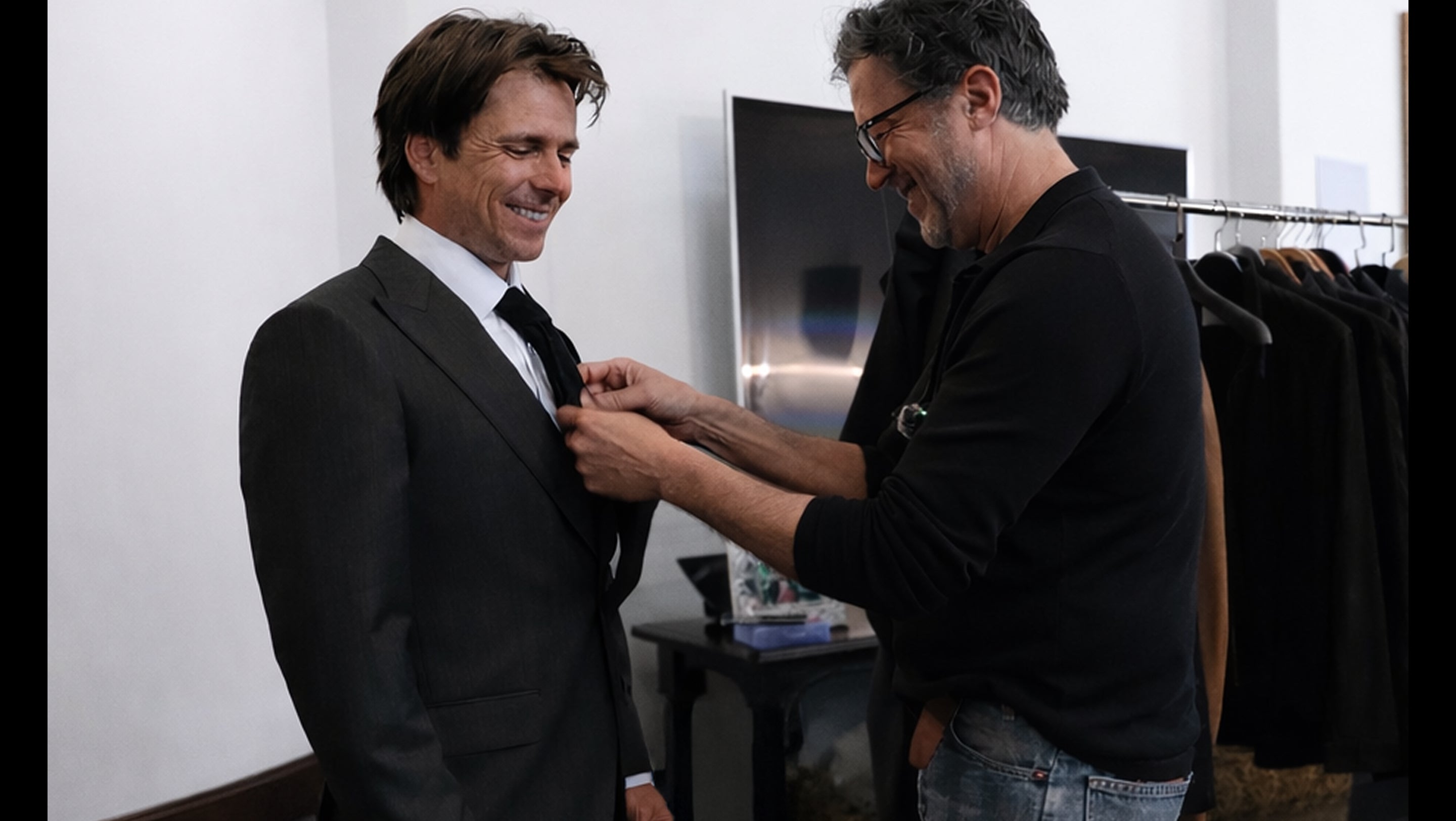 Man adjusting a suit on another man in a room with a television in the background.