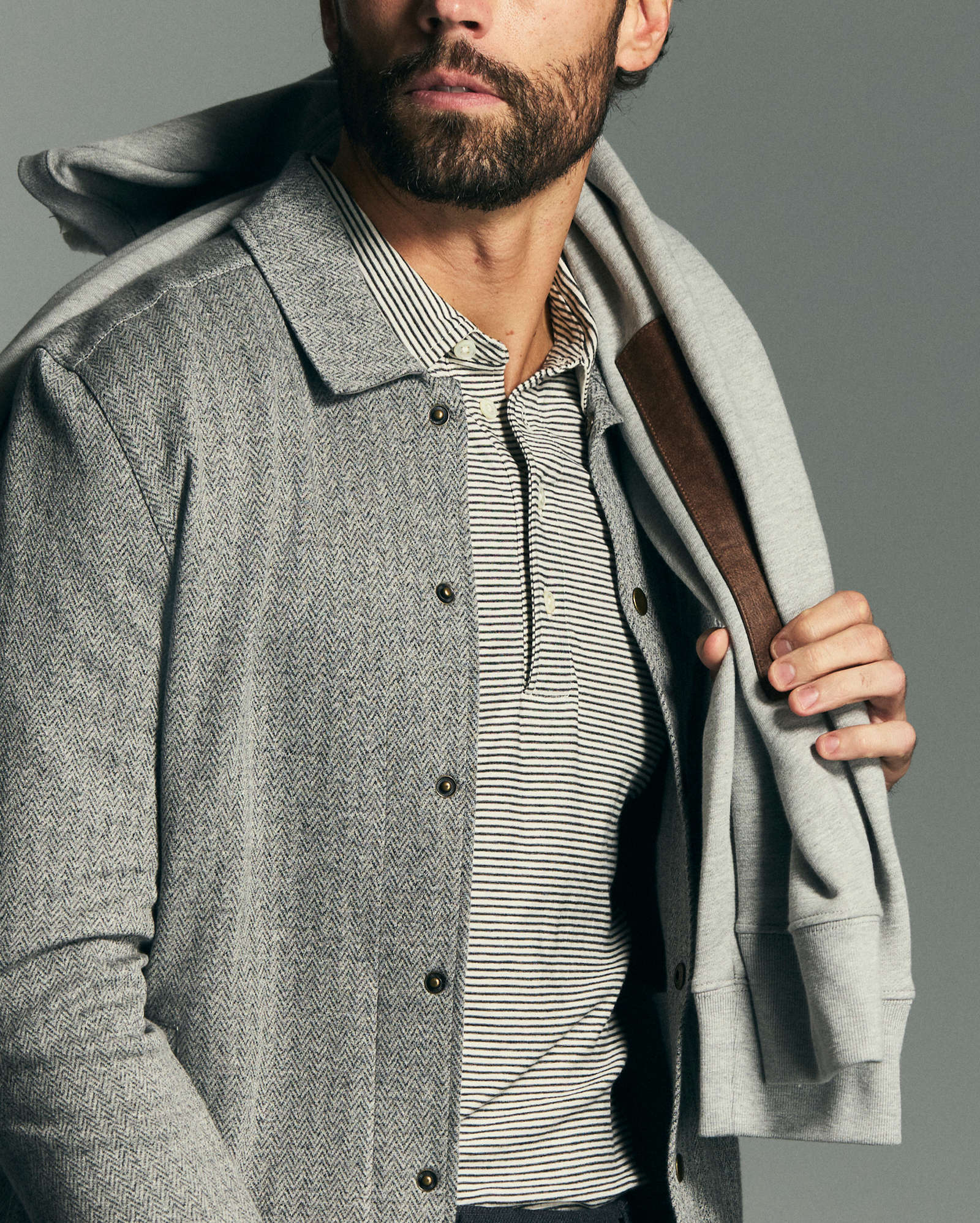 Man wearing a gray coat over a striped shirt with a brown leather bag over his shoulder on a gray background