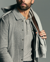 Man wearing a gray coat over a striped shirt with a brown leather bag over his shoulder on a gray background