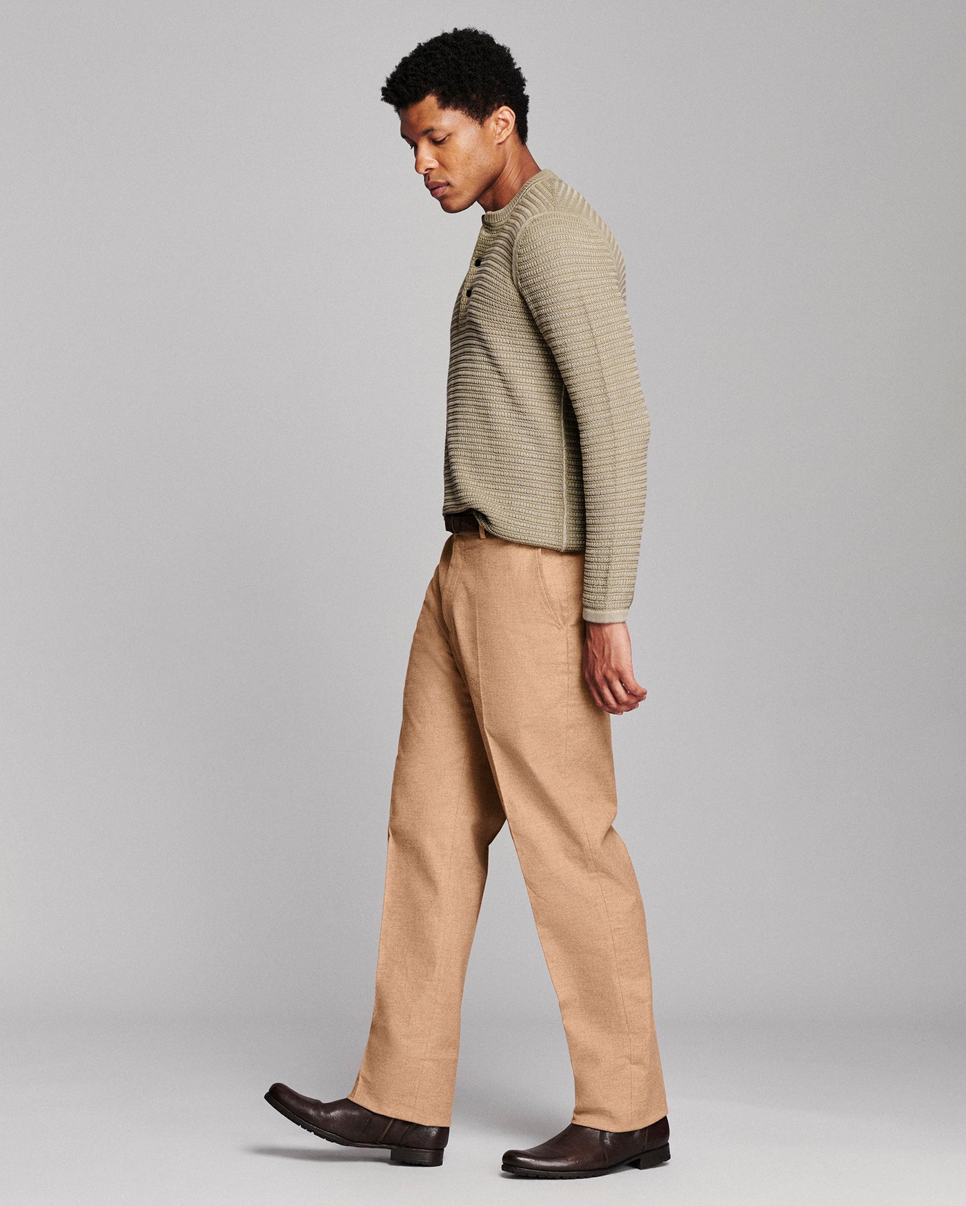 A man wearing a ribbed knit long-sleeve sweater in a light olive-beige color and tan trousers, shown in profile against a neutral gray studio background.