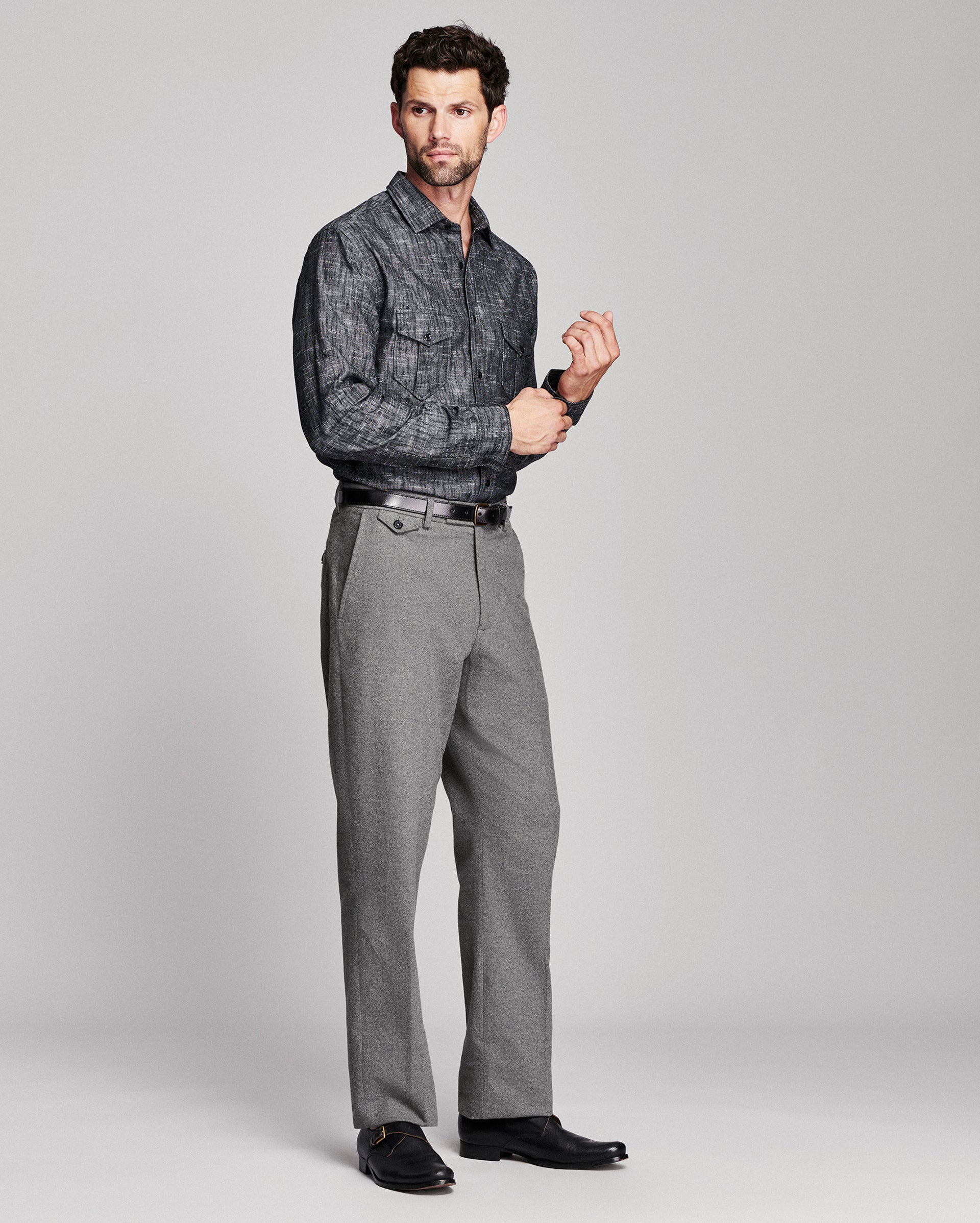 A man modeling a dark gray patterned long-sleeve button-down shirt with gray trousers, black belt, and black dress shoes, standing against a light gray studio background.