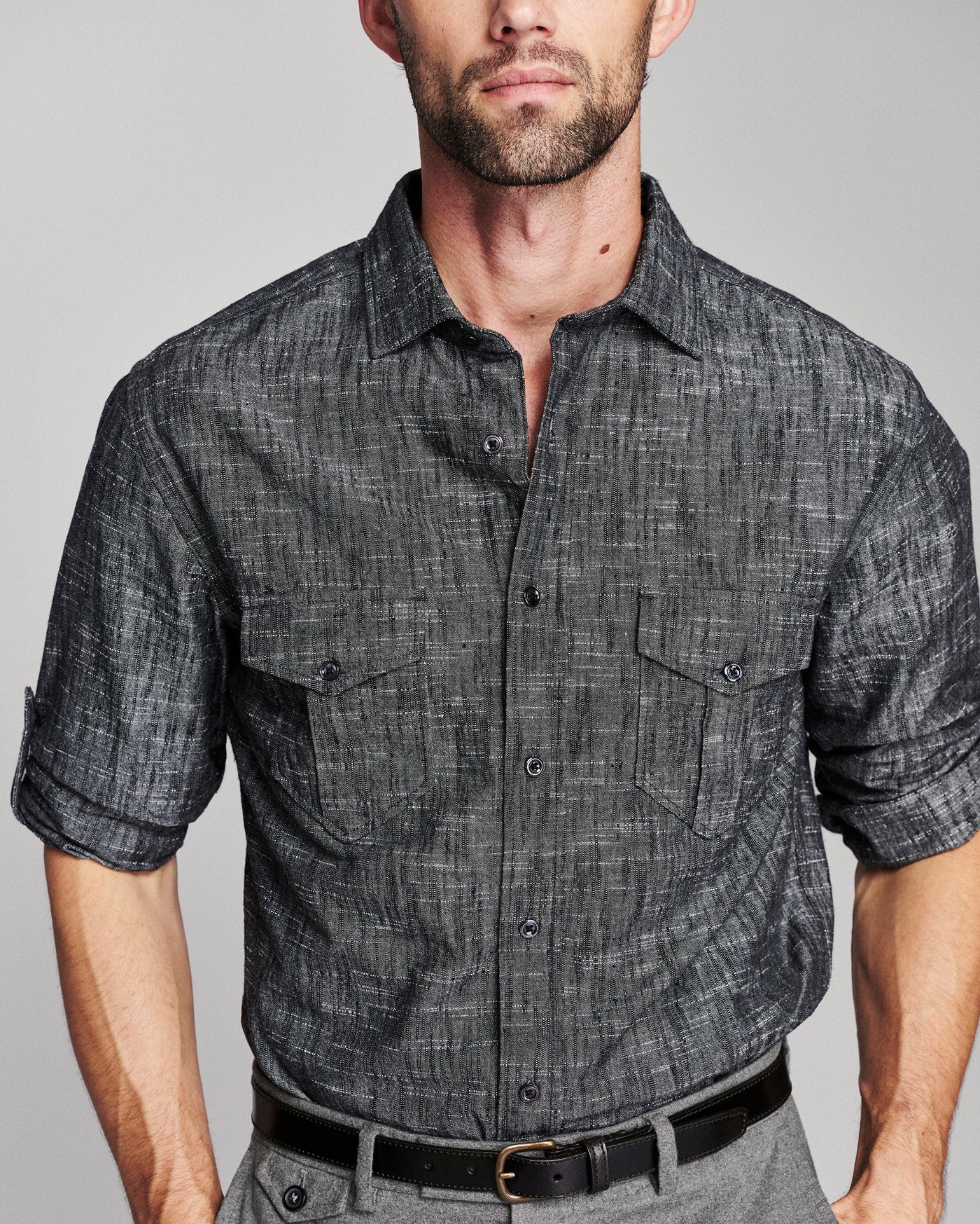 A man wearing a dark gray textured short-sleeve button-up shirt with two chest pockets, paired with gray trousers and a black belt, against a light gray studio background.