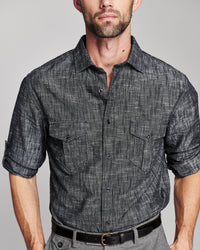 A man wearing a dark gray textured short-sleeve button-up shirt with two chest pockets, paired with gray trousers and a black belt, against a light gray studio background.