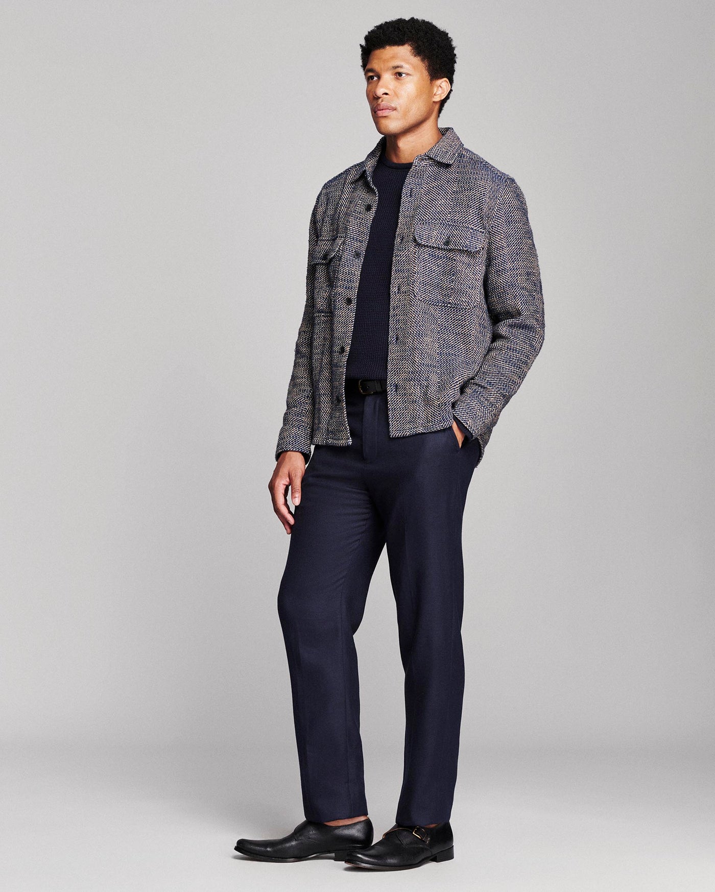 A man modeling a gray and navy tweed button-up shirt jacket over a dark navy t-shirt, paired with navy trousers and black leather dress shoes, against a light gray studio background.