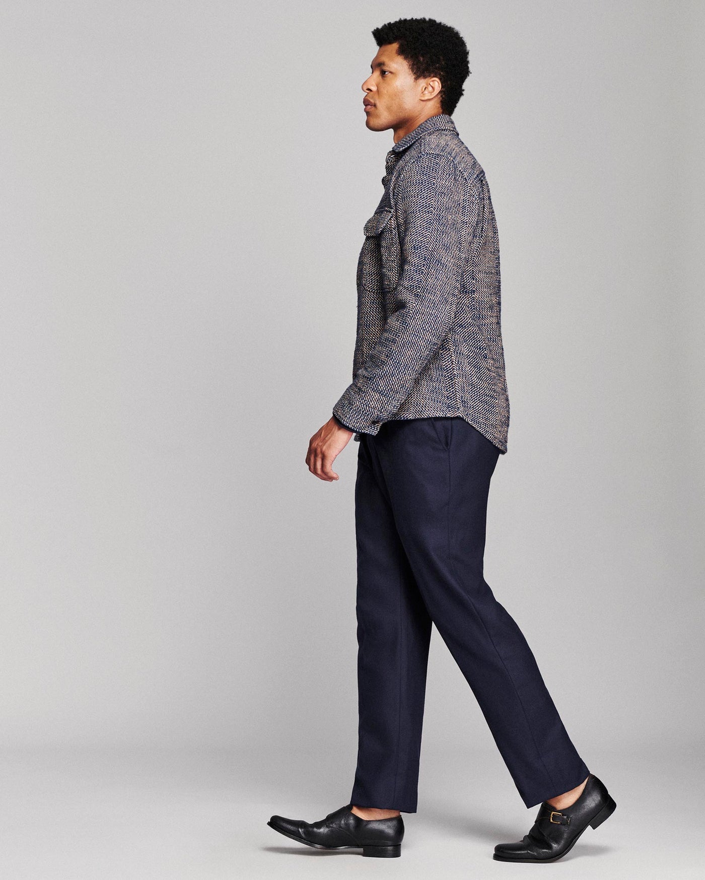 A man in side profile wearing a textured gray and blue patterned button-up shirt, navy blue trousers, and black leather dress shoes, photographed against a light gray studio background.