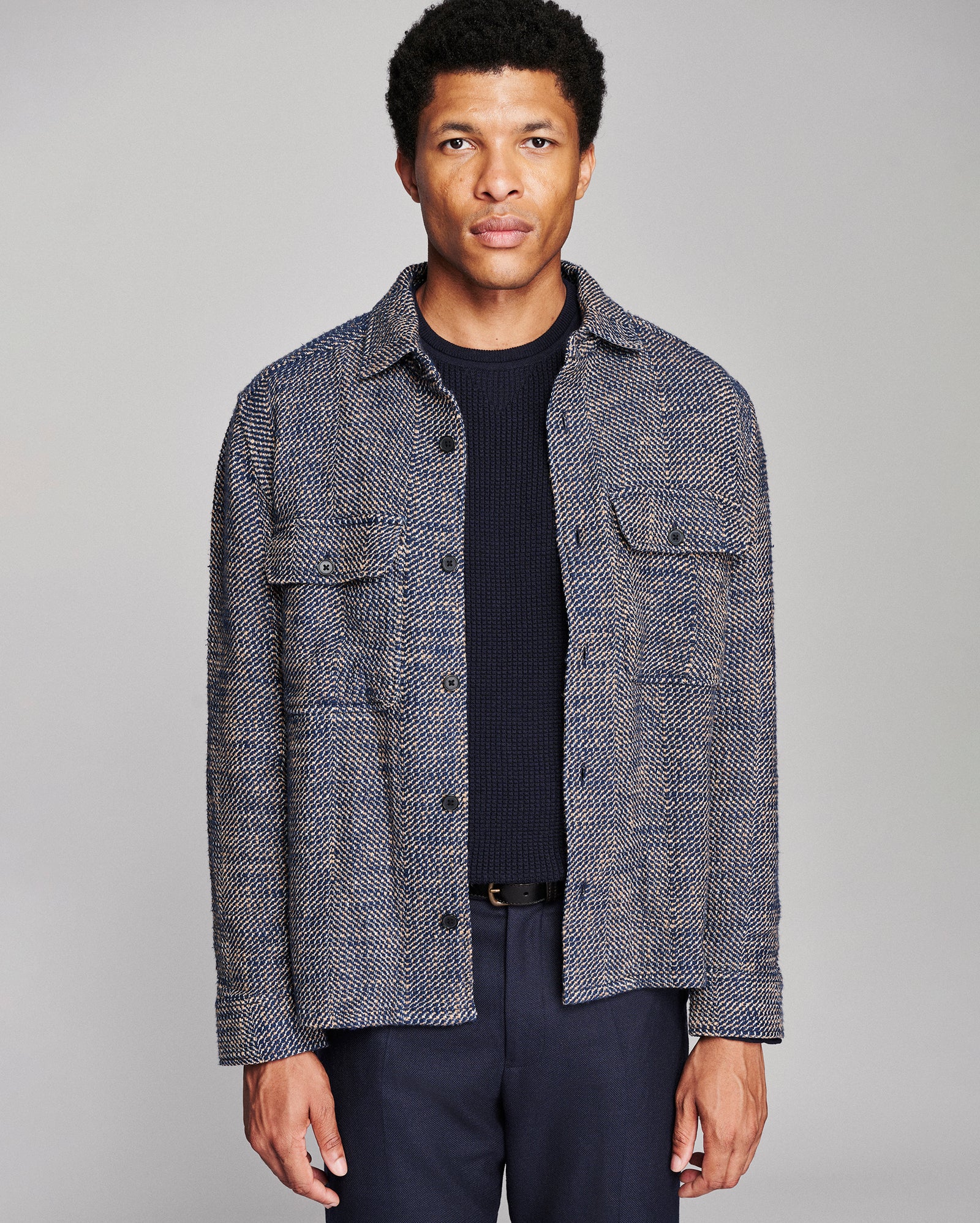 A man wearing a textured gray and navy tweed button-up shirt jacket over a dark navy ribbed sweater and navy trousers, photographed against a solid light gray studio background.