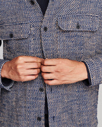 Close-up of a person wearing a textured herringbone tweed jacket, buttoning the front. The jacket features a blue, gray, and white woven pattern with dark buttons and flap chest pockets.