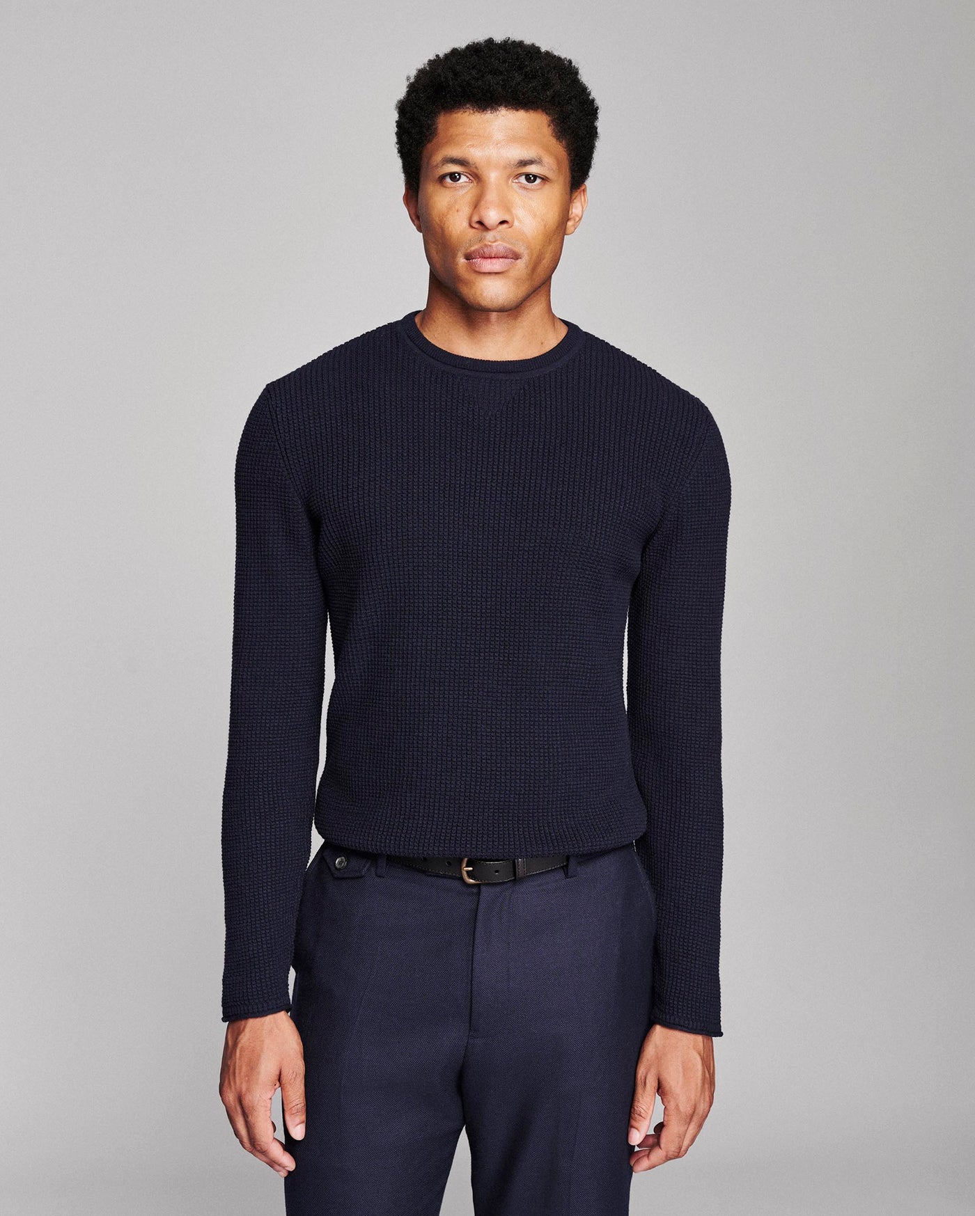 A man wearing a dark navy ribbed knit crewneck sweater and matching navy trousers, standing against a neutral gray studio background.