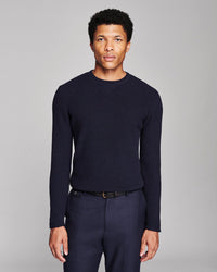 A man wearing a dark navy ribbed knit crewneck sweater and matching navy trousers, standing against a neutral gray studio background.