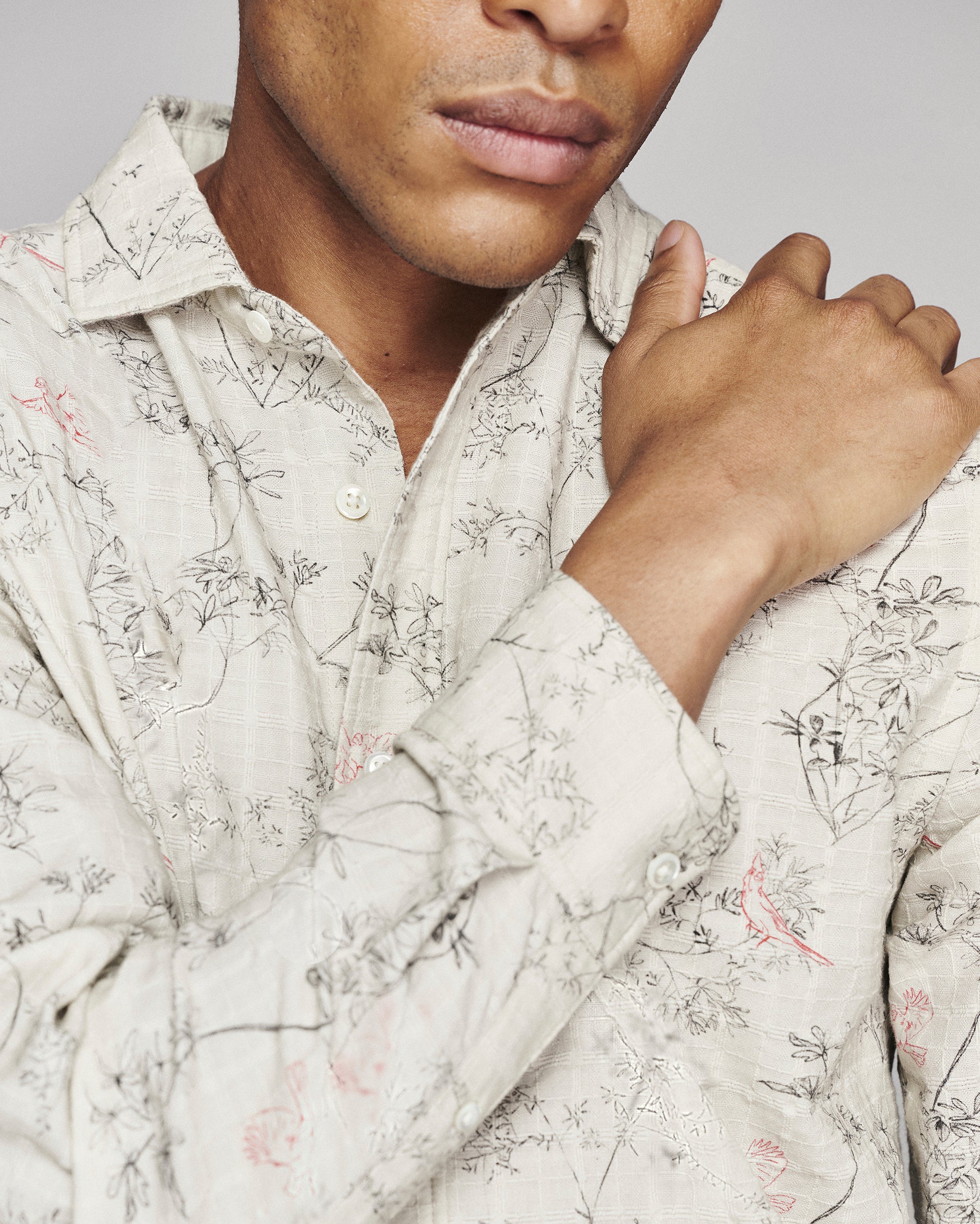 A close-up of a man wearing a light beige long-sleeve button-up shirt with a delicate black and pink botanical pattern, adjusting the collar with one hand.