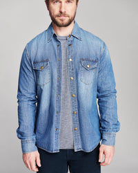 A man wearing a light blue denim button-up shirt over a gray striped t-shirt and dark pants, photographed against a solid light gray studio background.