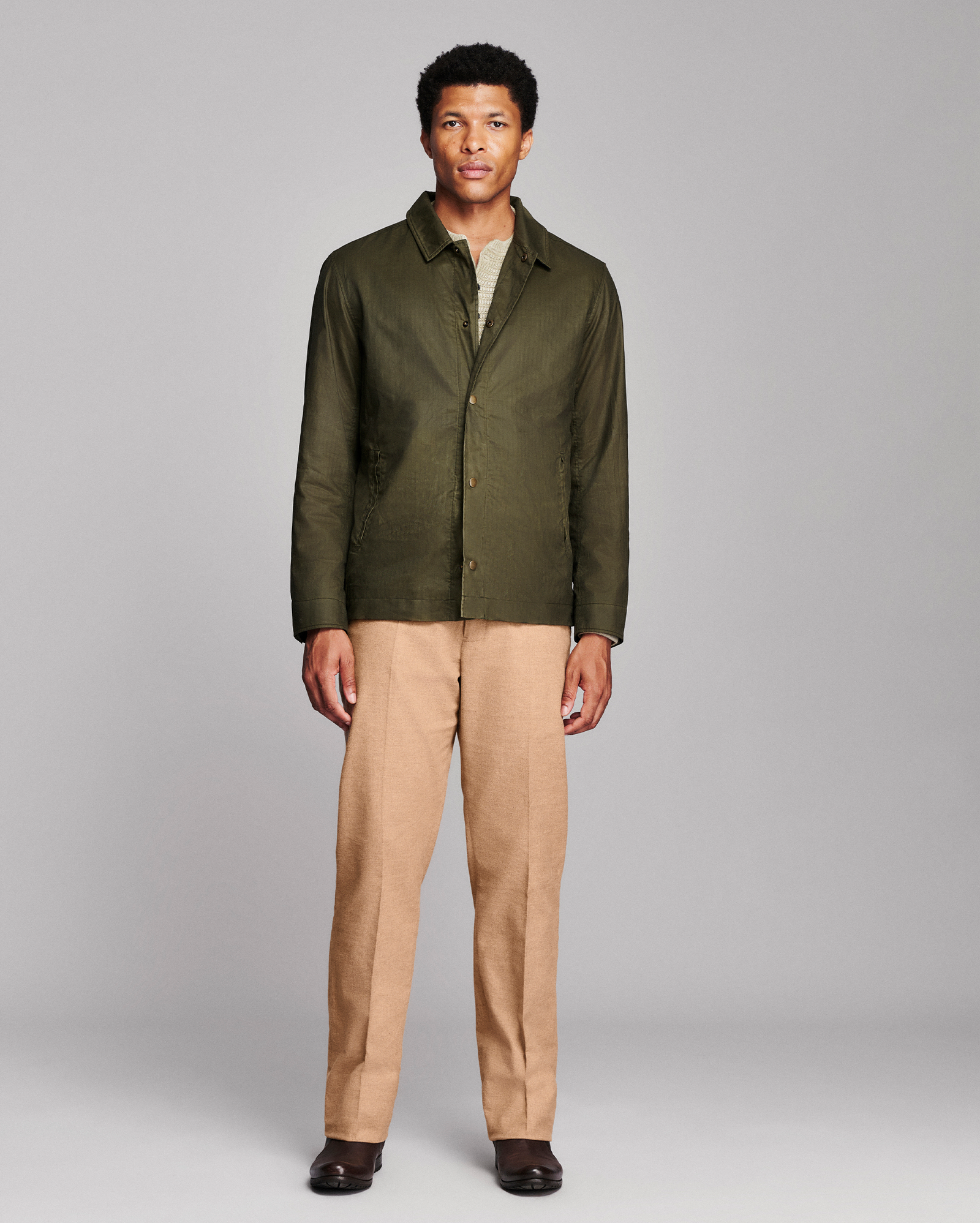 A man modeling an olive green button-up shirt jacket over a light-colored undershirt, paired with tan trousers and dark brown shoes, against a solid gray studio background.
