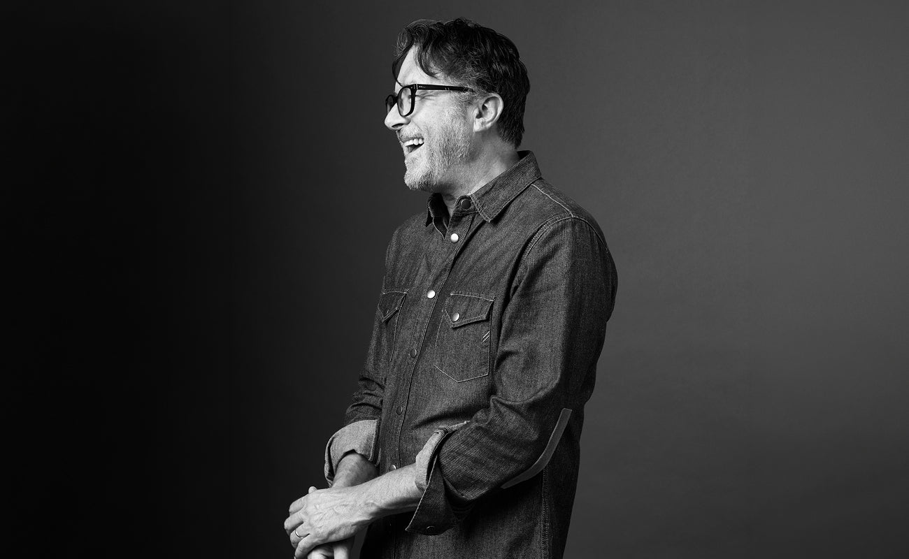 Billy Reid wears a denim shirt while laughing in a black and white photo.
