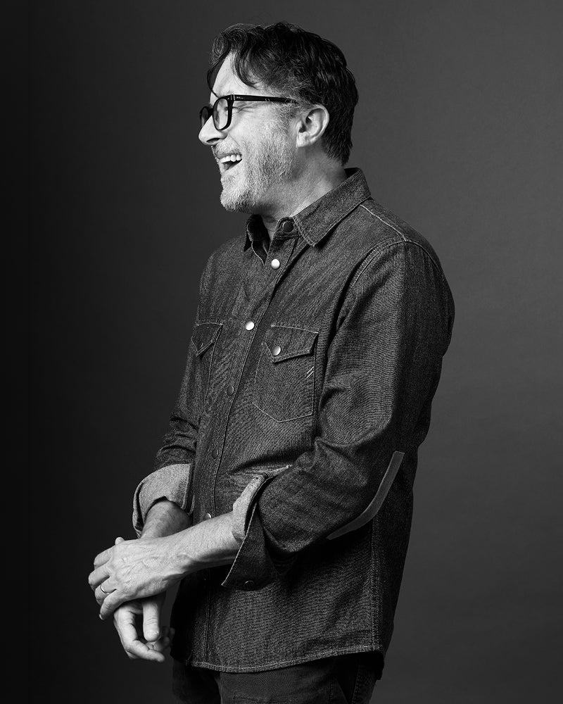 Billy Reid wears a denim shirt while laughing in a black and white photo.