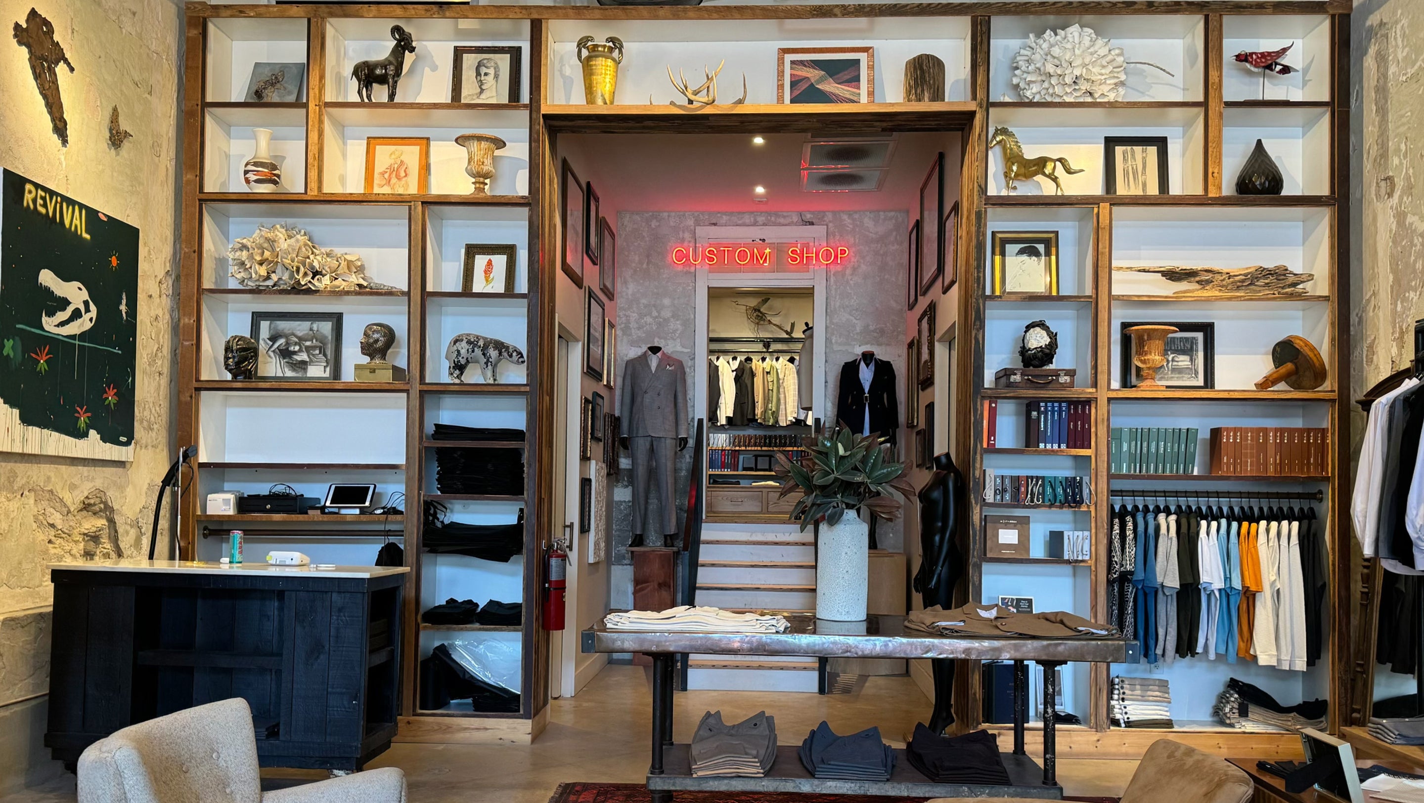 A boutique interior with tall wooden shelving units displaying framed art, decorative objects, books, and clothing racks. A central doorway leads to a staircase and more apparel, with a table and chairs in the foreground.