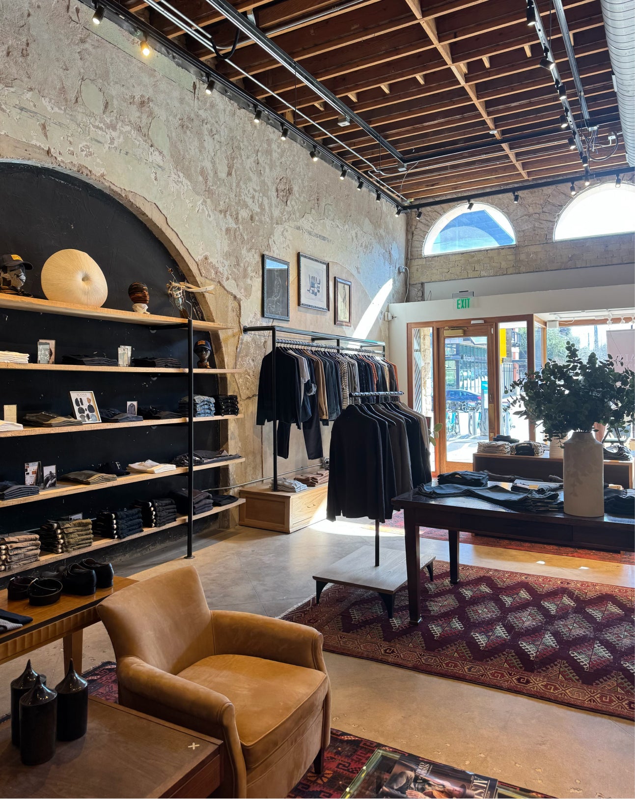 A boutique clothing store interior with racks of dark shirts, wooden shelving, a tan leather armchair, patterned rugs, and a high ceiling with exposed wood beams and track lighting.
