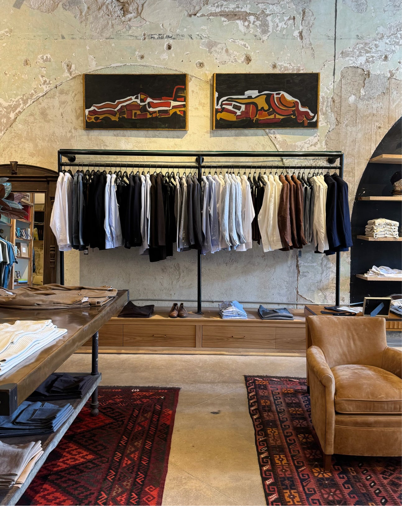 A retail clothing store interior with a black clothing rack holding multiple shirts and sweaters, two framed abstract paintings on a textured plaster wall, a leather armchair, patterned rugs, and display tables in the foreground.