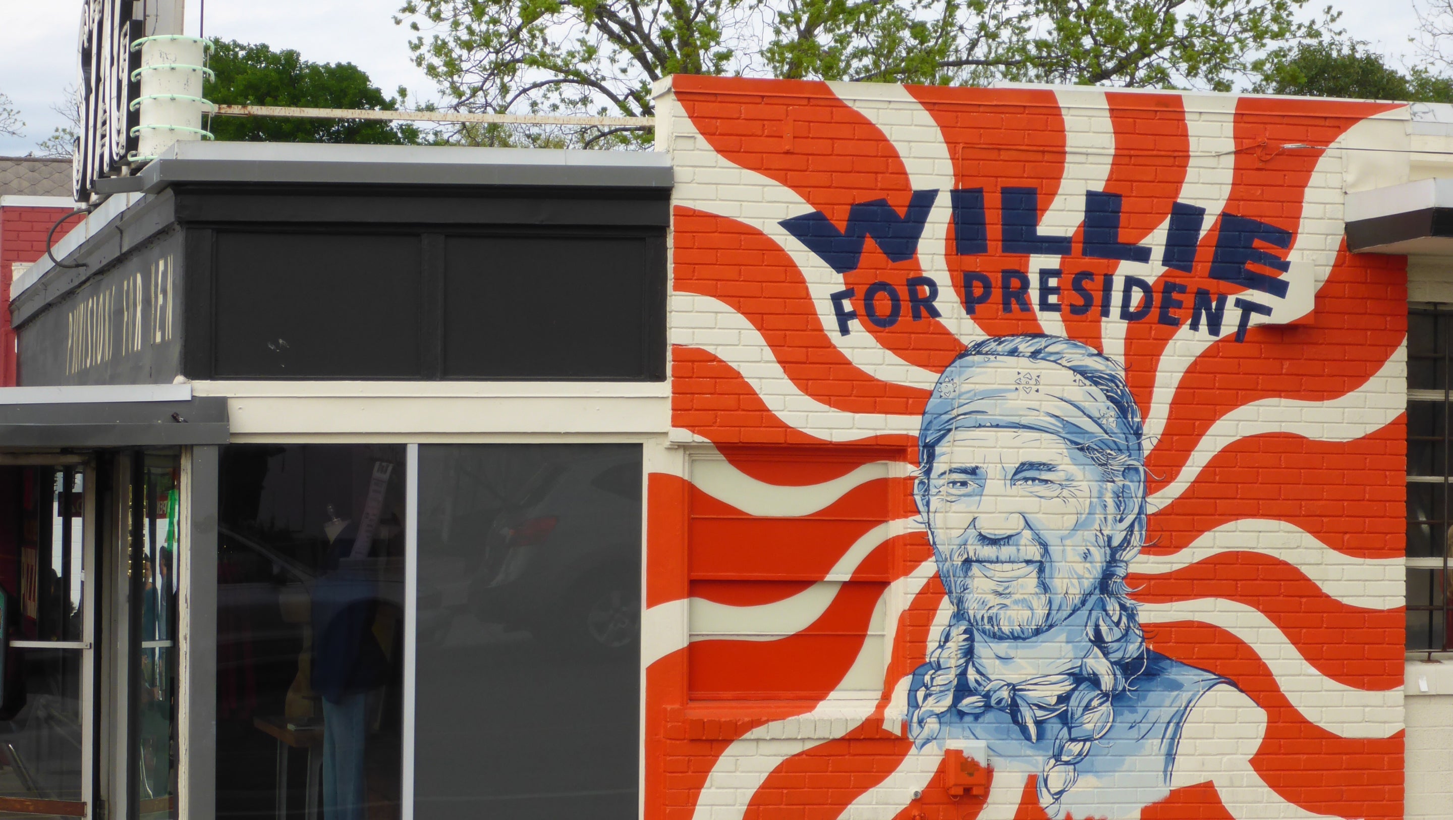 A large painted mural on a building wall reads “WILLIE FOR PRESIDENT” in blue text over a red and white wavy sunburst design, with a blue-and-white portrait of Willie Nelson. Part of a dark storefront and trees are visible at the left and top edges.