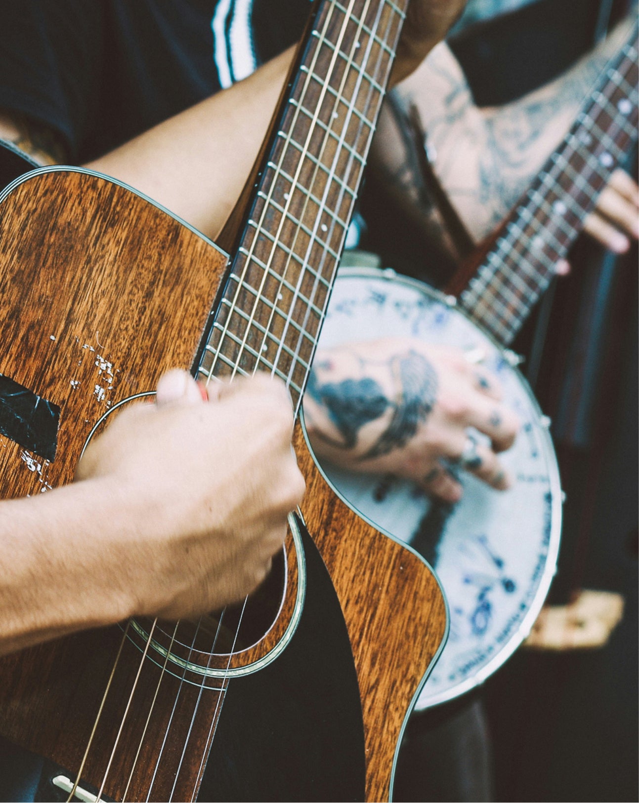 Close-up of a person playing an acoustic guitar, with tattooed hands visible on the neck and strumming, while another person holds a banjo in a dimly lit setting.
