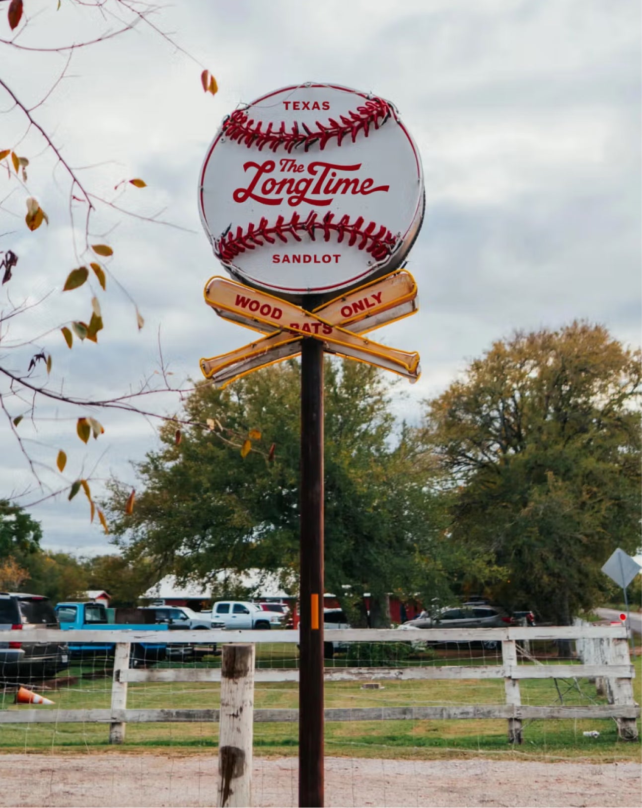 A decorative outdoor signpost featuring a baseball-shaped sign that reads 'The Longtime Texas Sandlot' with crossed wooden baseball-club signs below reading 'Wood Only' and 'Only Wood'. The sign is photographed outdoors in a rural setting with trees, a fence, and vehicles in the background.