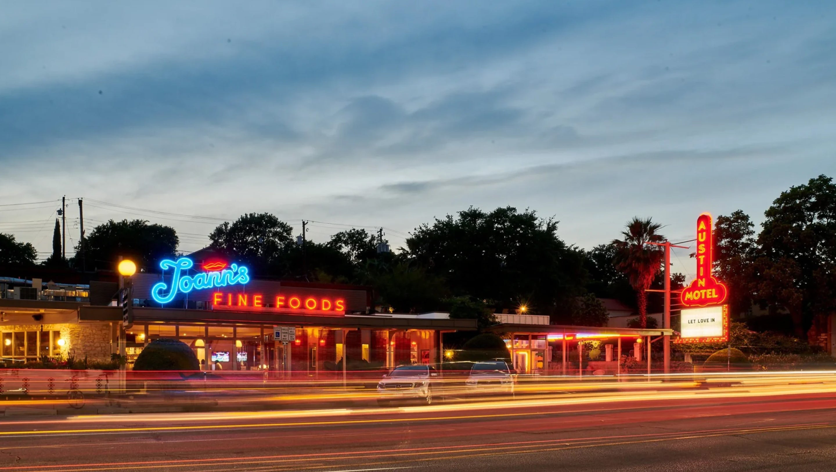 A nighttime street scene featuring a retro motel and diner with glowing neon signs, including a prominent blue 'Joan's Fine Foods' sign and a red motel sign. Long-exposure light trails from passing cars stretch across the foreground, with trees and a cloudy dusk sky in the background.