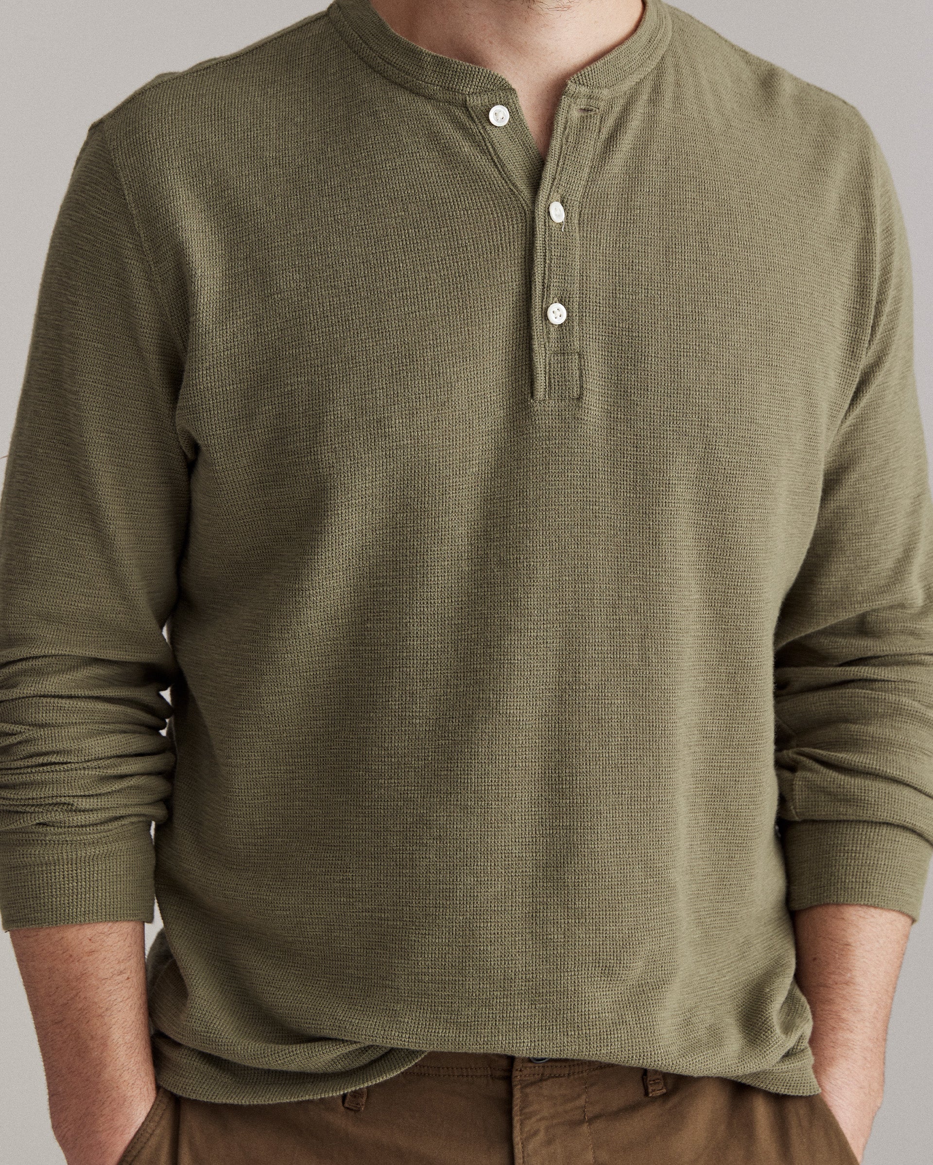 A person wearing an olive green, long-sleeve Henley shirt with three white buttons, and brown pants. The persons face is partially out of frame.