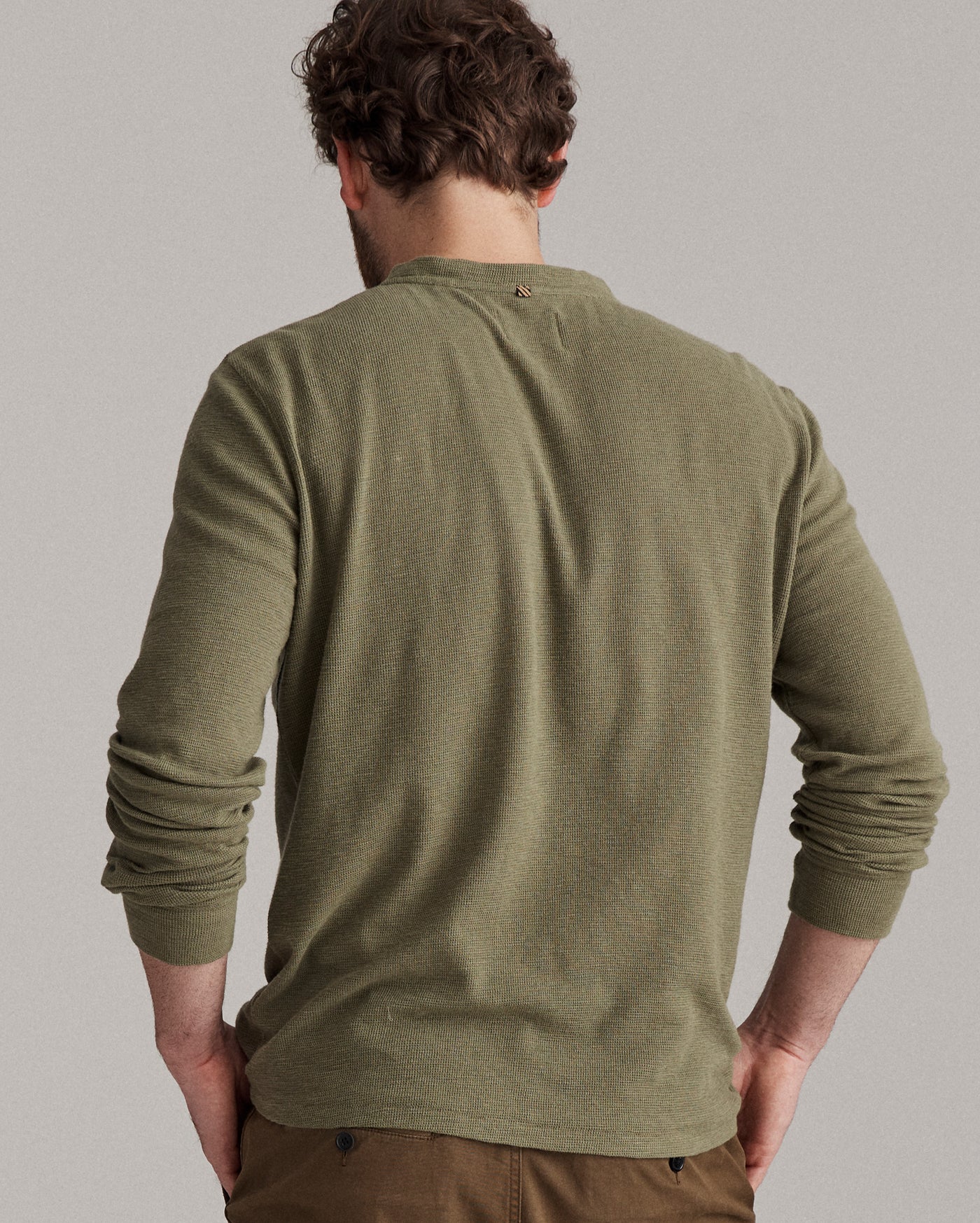 A man with curly brown hair stands facing away, wearing an olive green long-sleeve shirt and brown pants. His hands rest on his hips against a plain gray background.