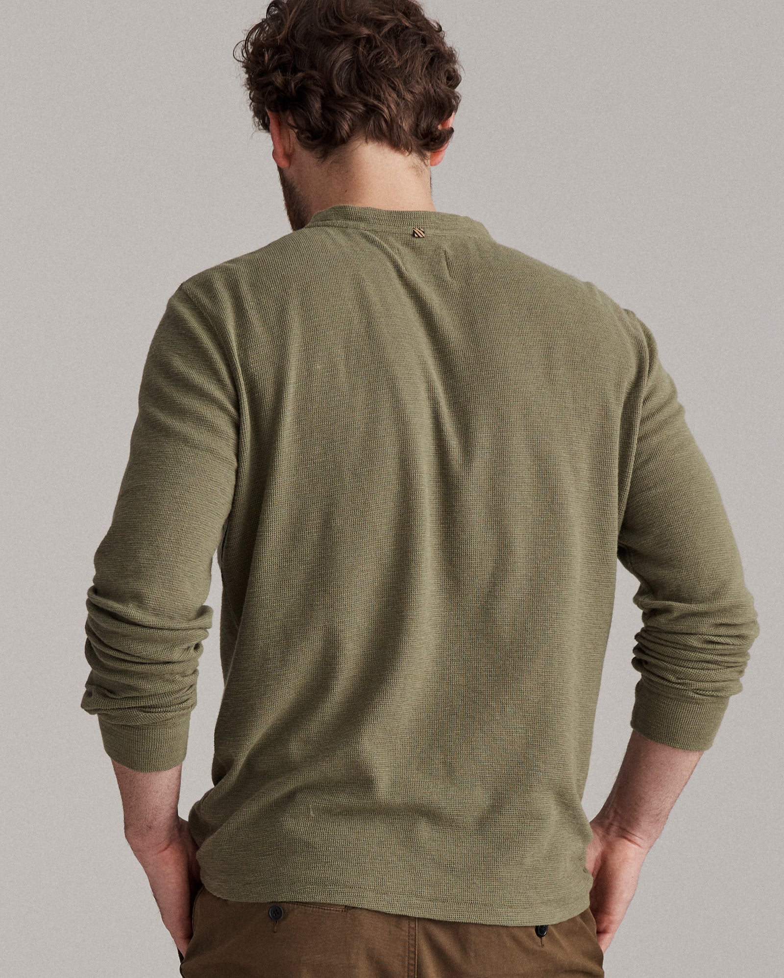A man with curly brown hair stands facing away, wearing an olive green long-sleeve shirt and brown pants. His hands rest on his hips against a plain gray background.