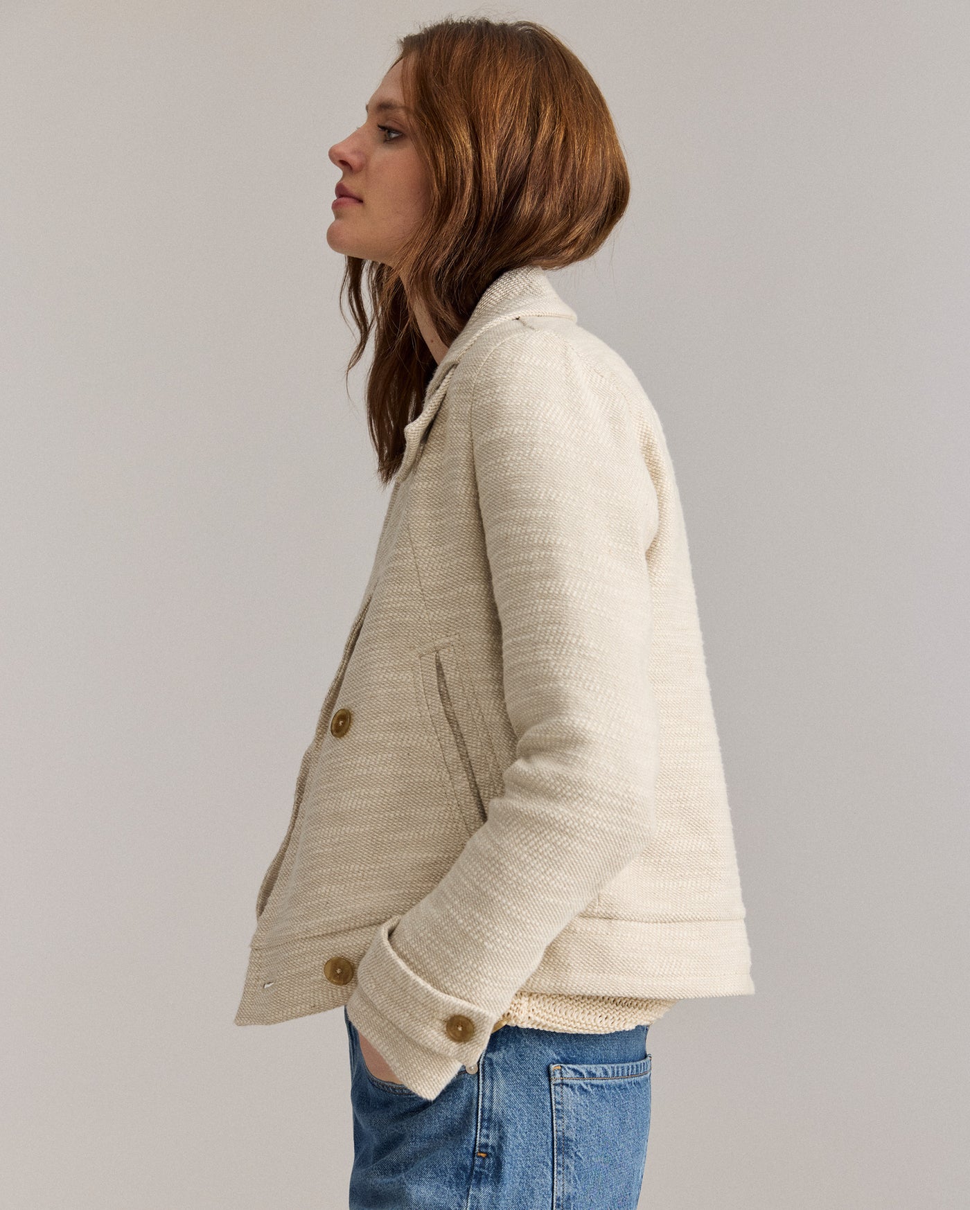 A woman with long brown hair wears a cream-colored textured jacket and blue jeans, standing in profile with her left hand in her pocket against a plain light background.
