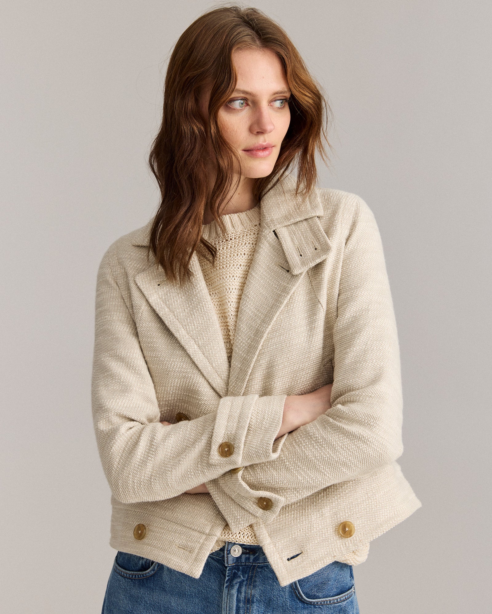 A woman with wavy brown hair, wearing a light beige textured jacket over a knit sweater and blue jeans, stands with her arms folded and looks to the side against a plain background.