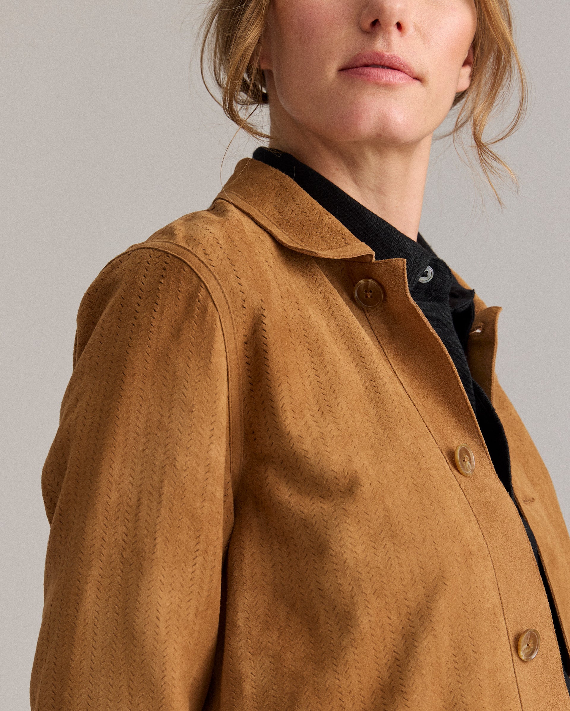 A woman wearing a light brown button-up jacket over a black top, pictured from the shoulders up against a plain background. Only the lower part of her face and upper torso are visible.
