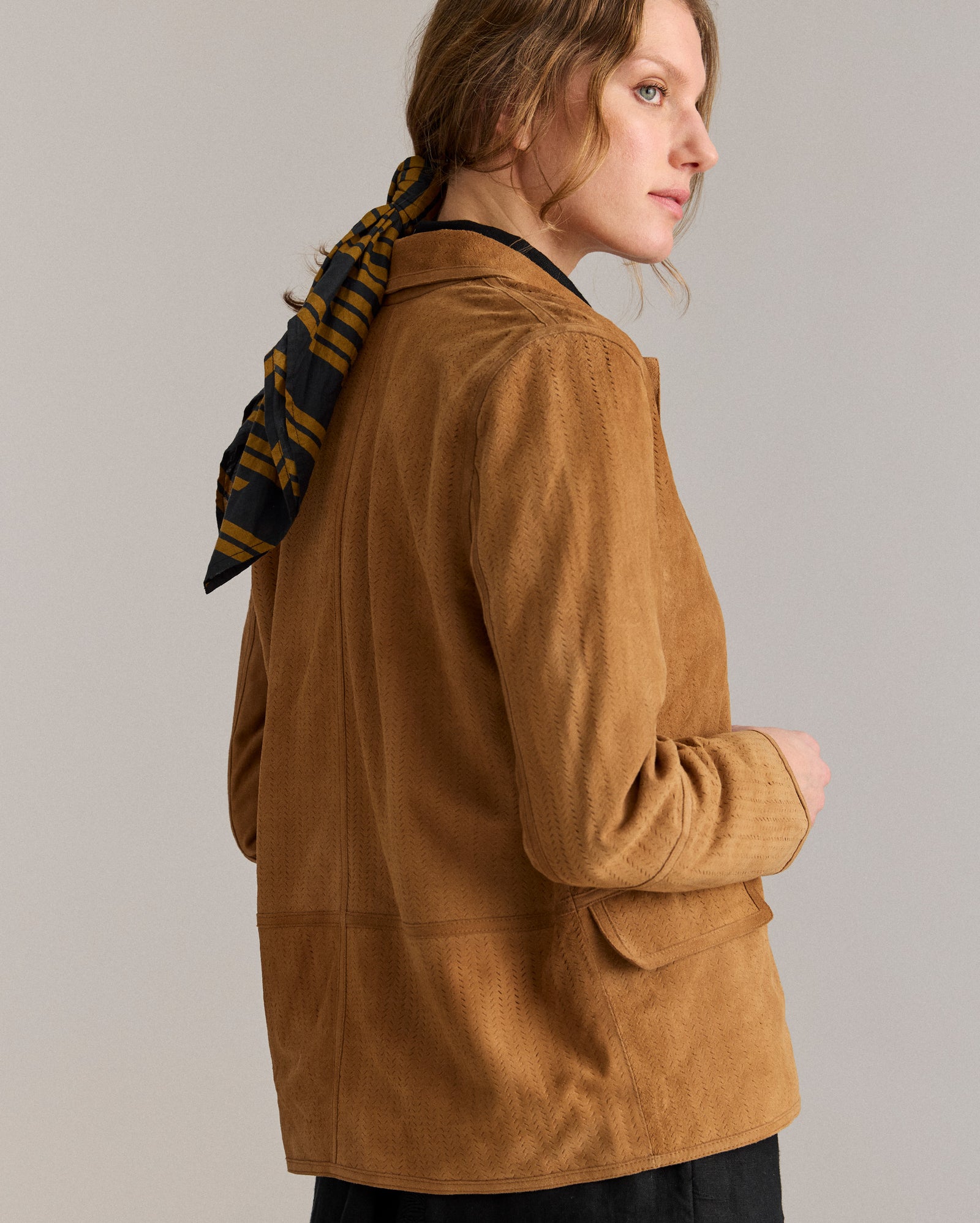 A person with wavy hair tied back with a black and brown striped scarf is wearing a tan suede jacket and black pants, standing sideways and looking over their shoulder against a plain background.