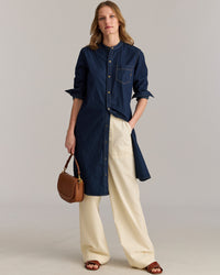 A woman stands against a plain background wearing a dark denim button-down tunic over cream wide-leg pants, brown sandals, and holding a brown shoulder bag. Her sleeves are rolled up and she has loose, wavy hair.
