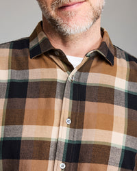 A close-up of a man wearing a brown, black, and beige plaid button-up shirt. His face is partially visible, showing a light beard and a subtle smile. The shirt has a collar and white buttons.
