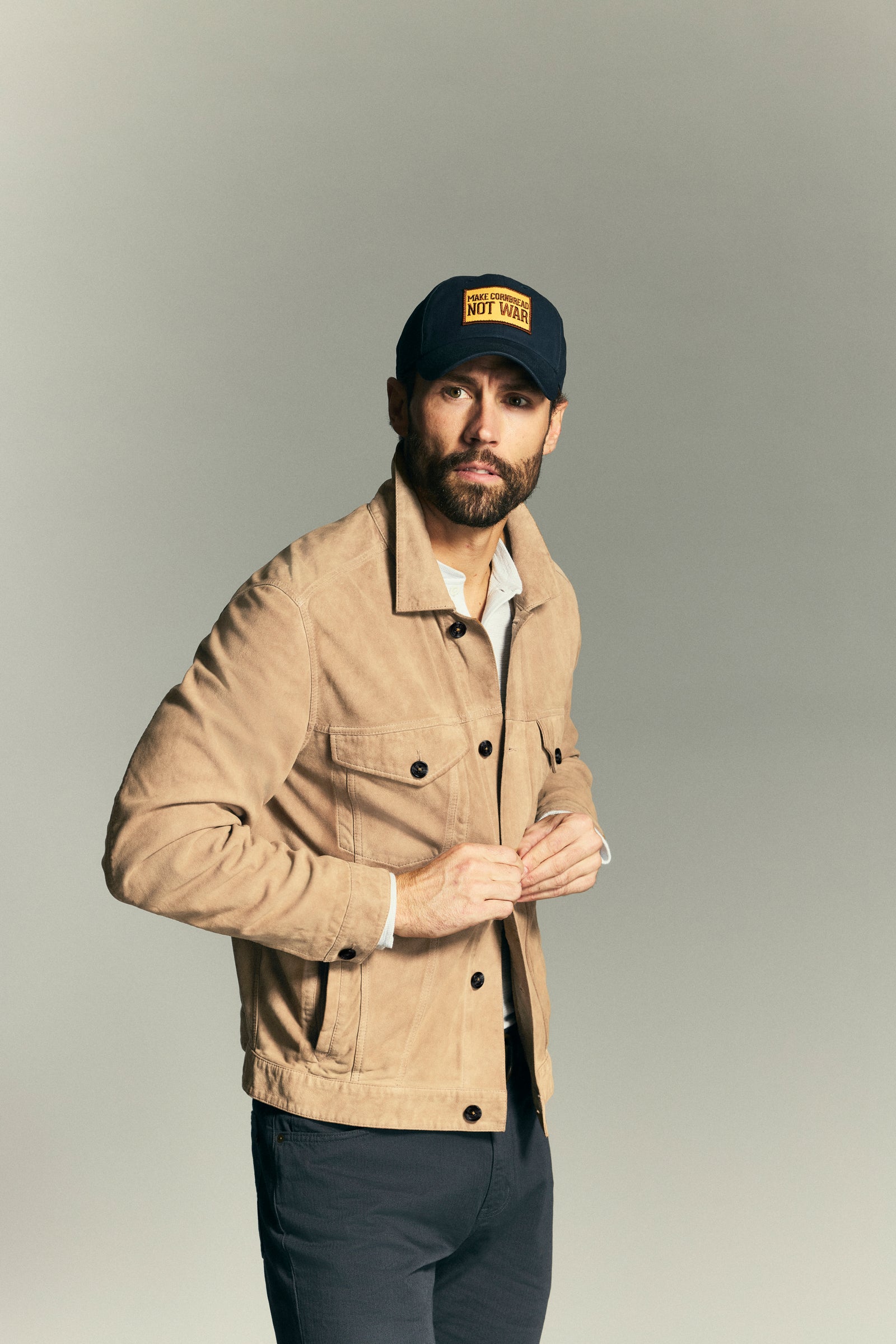 A man wearing a tan suede jacket, a dark navy baseball cap with a yellow logo patch, and dark trousers, posing against a neutral gray studio background.