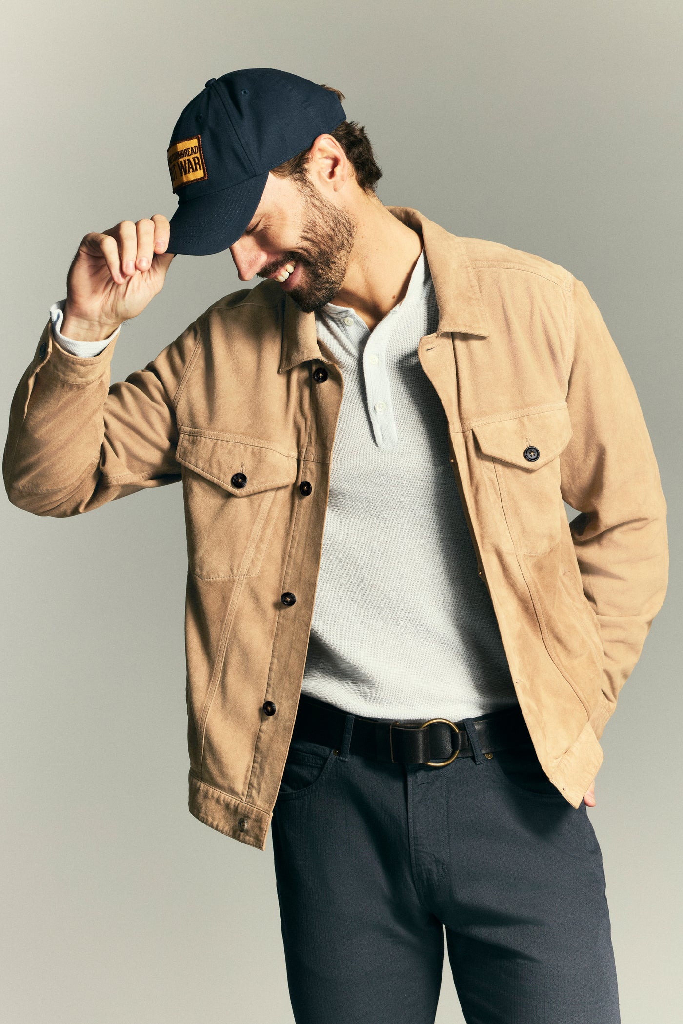 A man wearing a tan suede jacket over a light gray polo shirt, dark gray pants, and a navy blue baseball cap, posed against a neutral gray studio background.