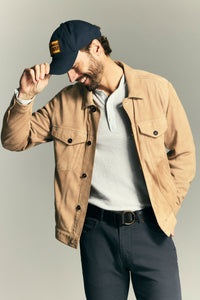 A man wearing a tan suede jacket over a light gray polo shirt, dark gray pants, and a navy blue baseball cap, posed against a neutral gray studio background.