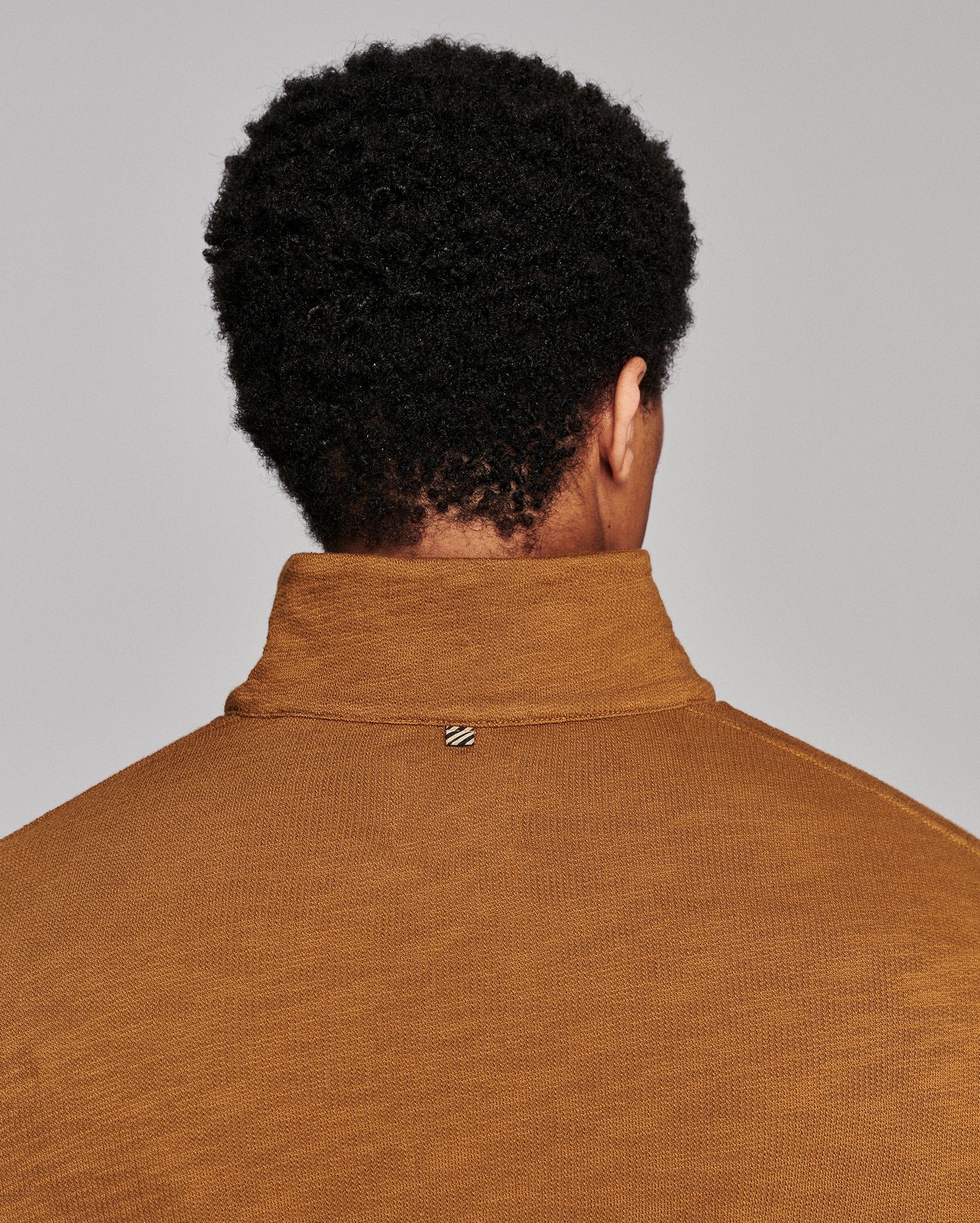 Back view of a person with curly black hair wearing a mustard brown ribbed knit mock neck sweater against a light gray background.