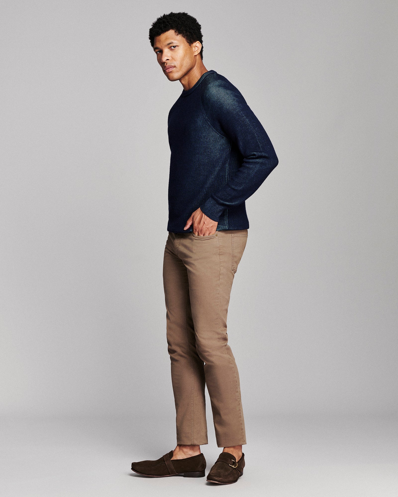 A man modeling a dark navy blue knit crewneck sweater, paired with tan trousers and dark brown loafers, against a solid light gray studio background.