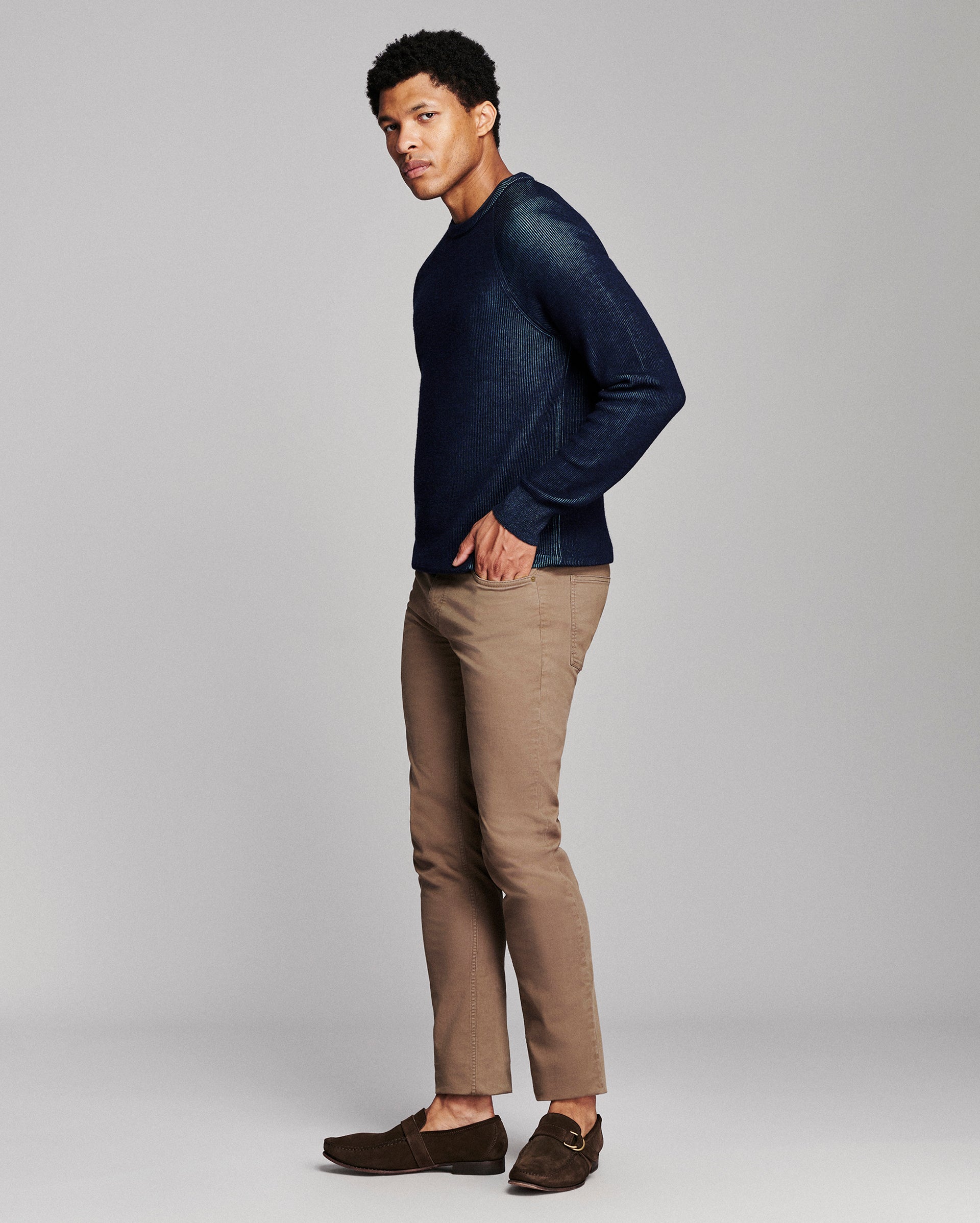 A man modeling a dark navy blue knit crewneck sweater, paired with tan trousers and dark brown loafers, against a solid light gray studio background.