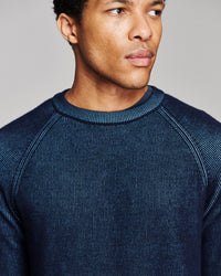 A close-up of a man wearing a dark navy blue ribbed knit crewneck sweater against a neutral gray background.