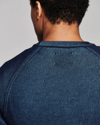 Close-up back view of a person wearing a dark blue ribbed knit crewneck sweater against a light gray studio background.