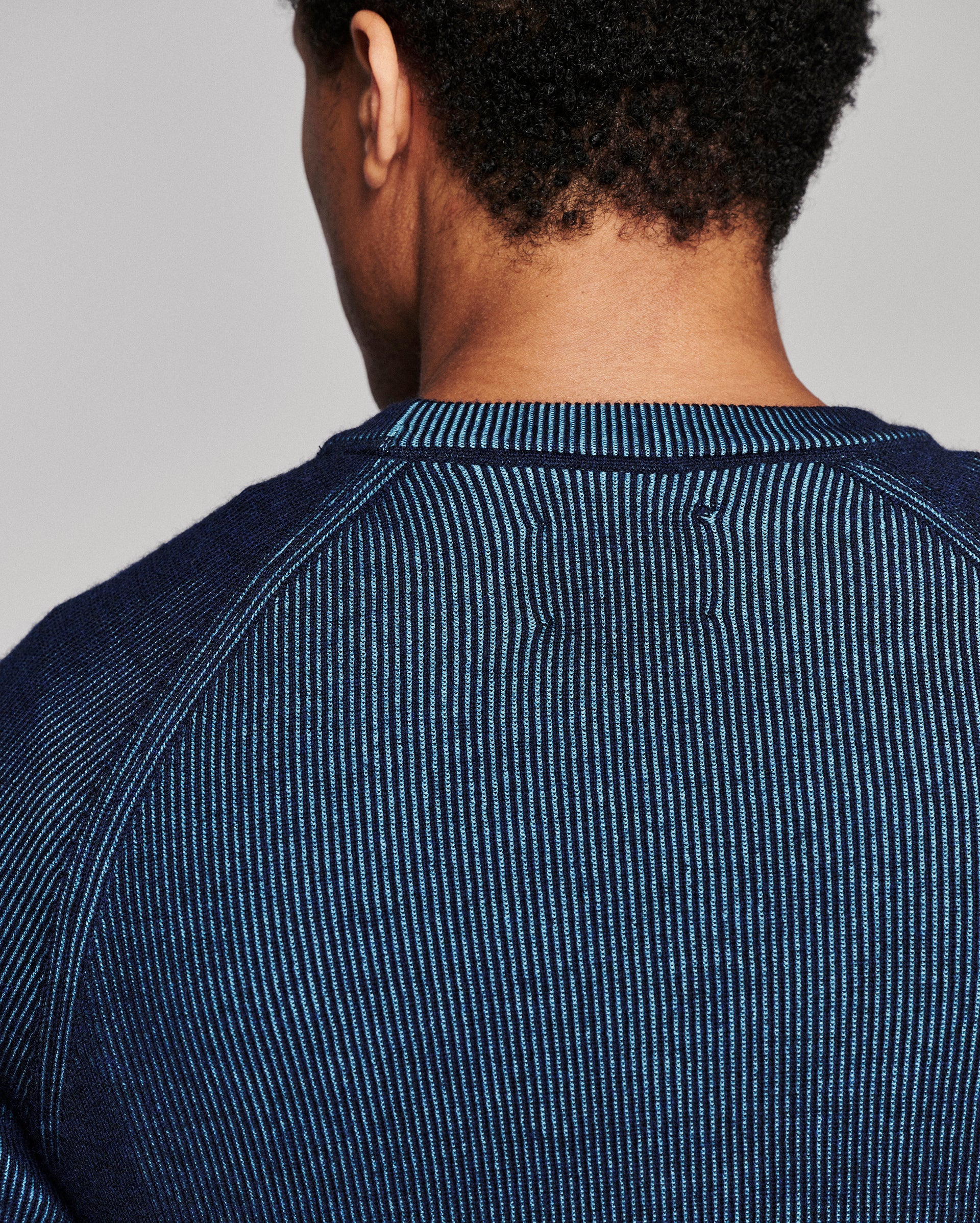 Close-up back view of a person wearing a dark blue ribbed knit crewneck sweater against a light gray studio background.