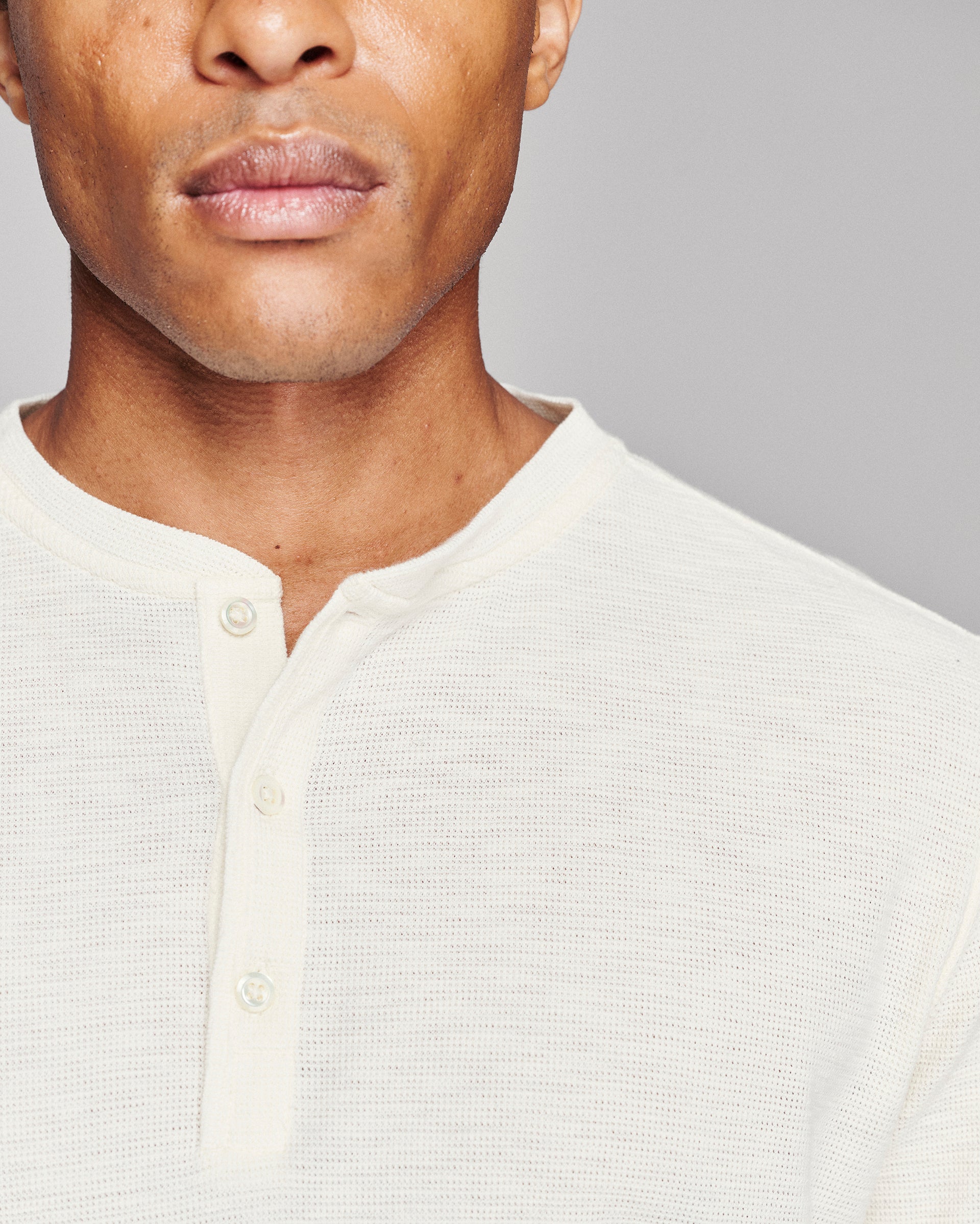 Close-up of a man wearing a textured off-white henley shirt against a plain gray background.