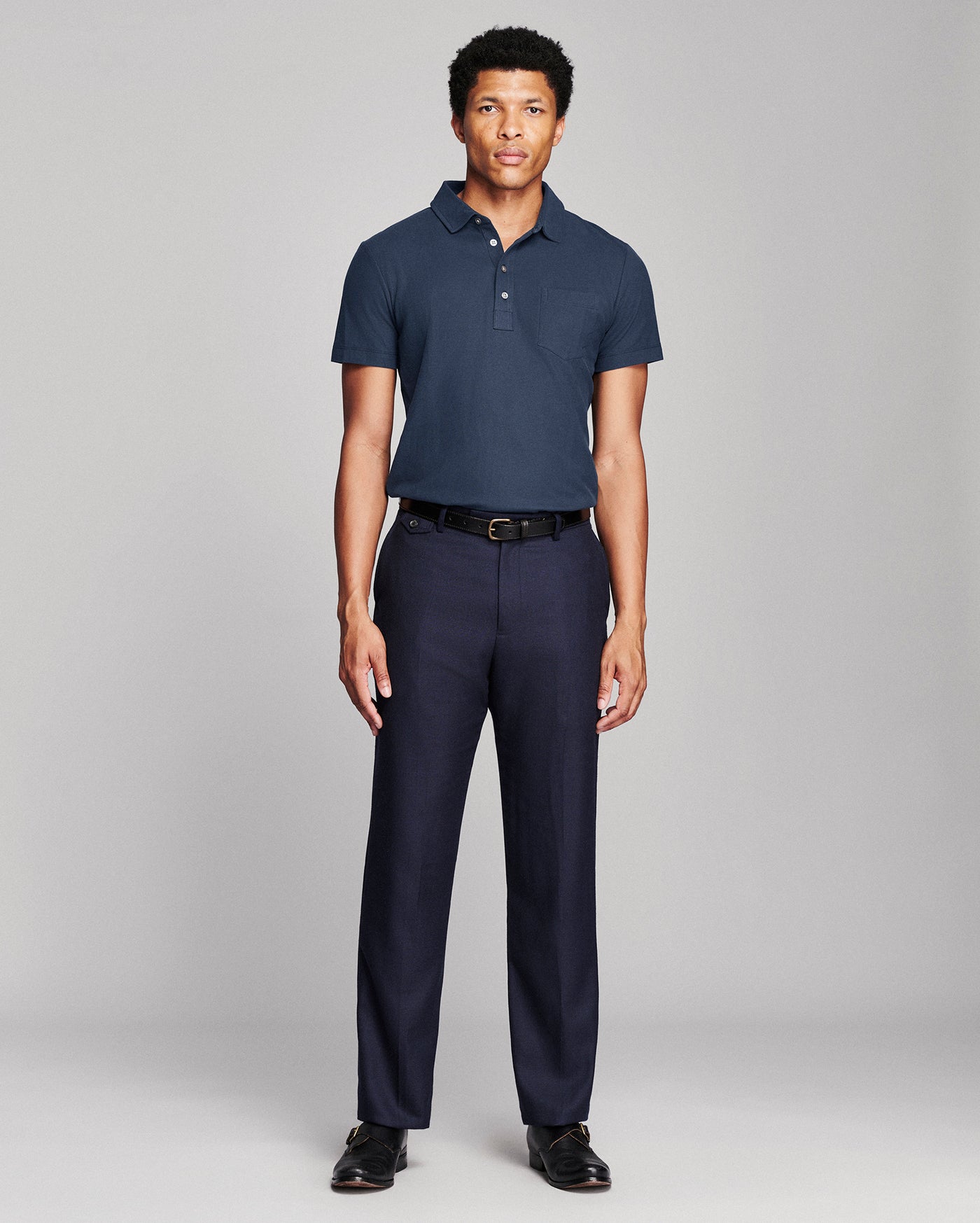 A man modeling a navy blue short-sleeve polo shirt and matching navy trousers with a black belt, standing against a light gray studio background.