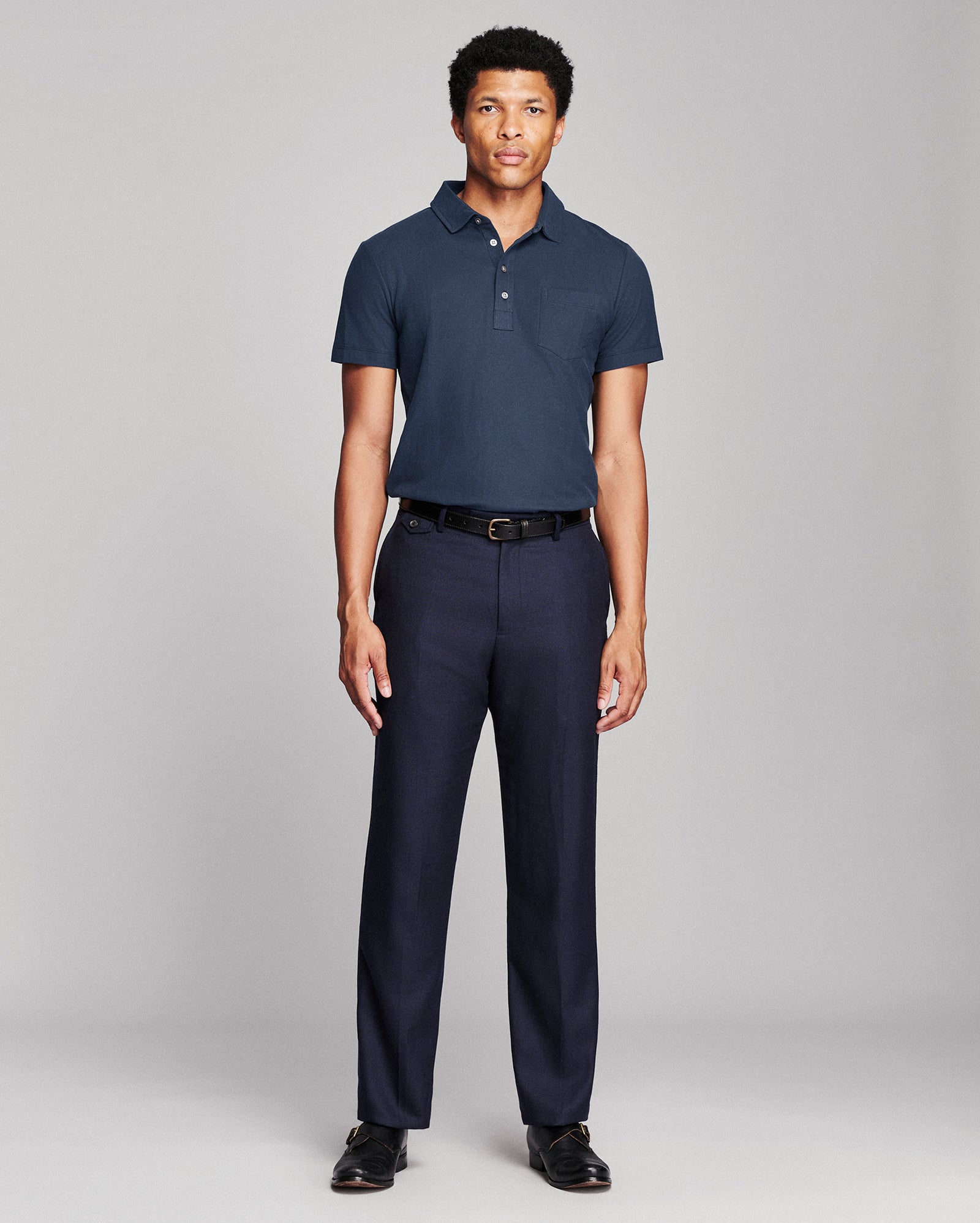A man modeling a navy blue short-sleeve polo shirt and matching navy trousers with a black belt, standing against a light gray studio background.