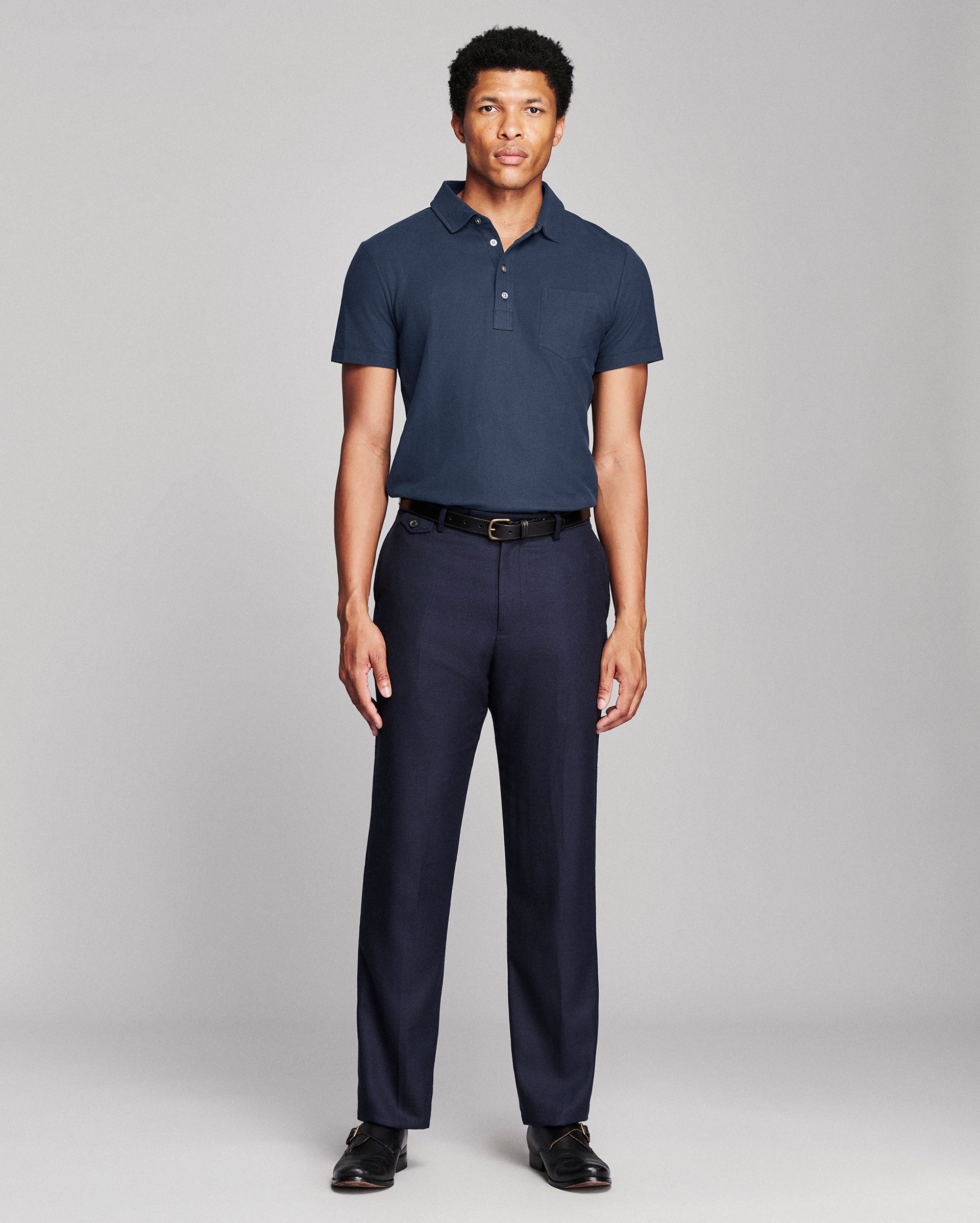 A man modeling a navy blue short-sleeve polo shirt and matching navy trousers with a black belt, standing against a light gray studio background.