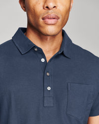 Close-up of a man wearing a dark navy blue short-sleeve polo shirt with a collar, three-button placket, and a chest pocket, set against a plain light gray background.