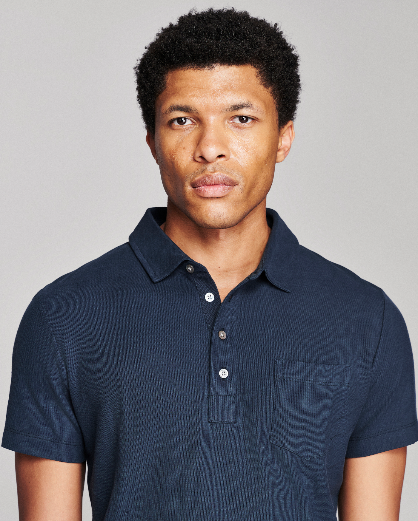 A man wearing a dark navy blue short-sleeve polo shirt with a three-button placket and a chest pocket, photographed against a solid light gray background.